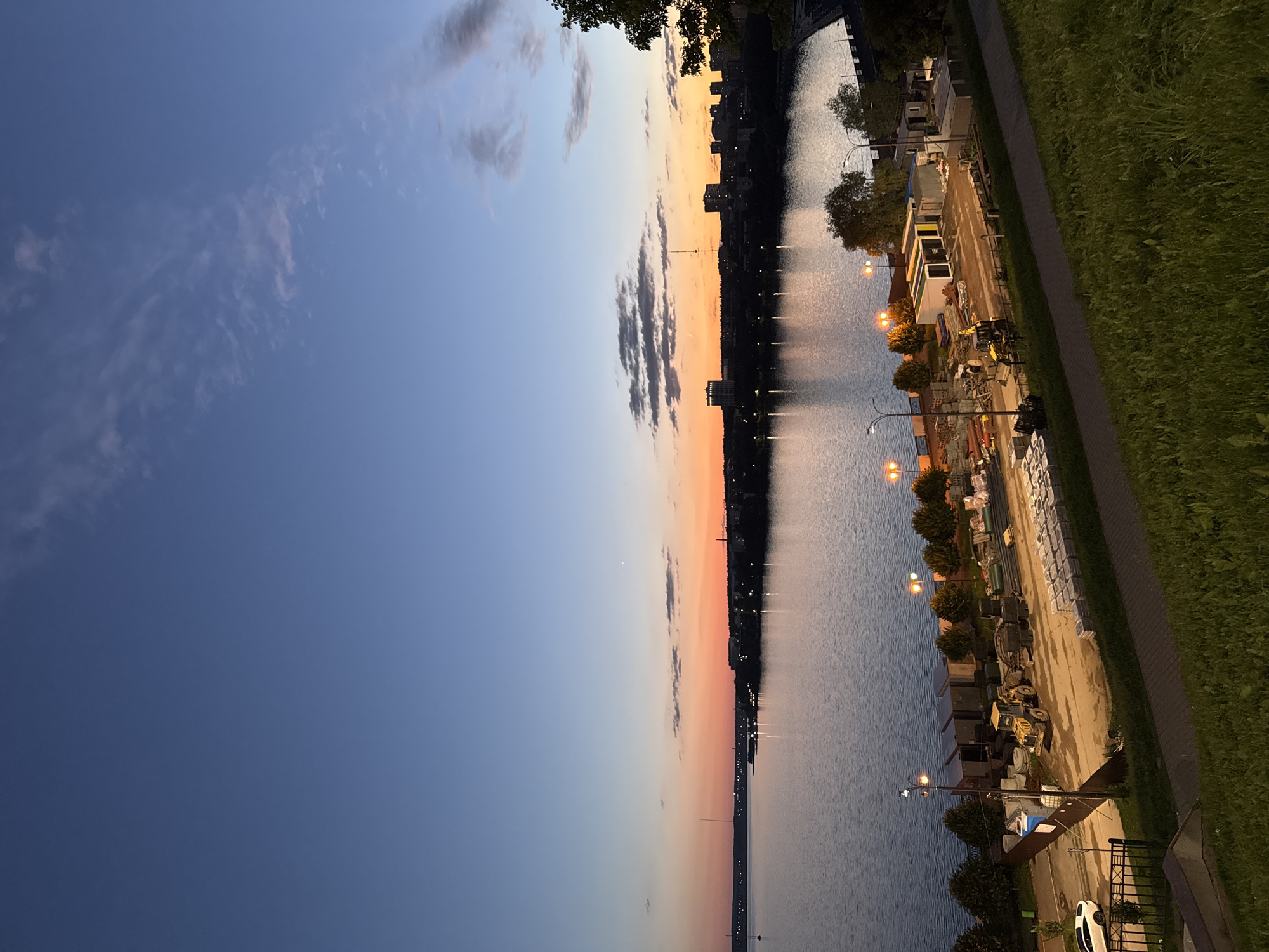 This is an outdoor, full-frame, eye-level, long shot photograph taken at dusk, overlooking a city skyline and a body of water, likely a river or a lake. The scene appears to be in Izhevsk, Russia, based on the architecture and the prominent Volga River.
The sky transitions from a deep blue at the top to a vibrant orange and pink sunset near the horizon. Wispy clouds are scattered across the sky. The city skyline is visible across the water, featuring a mix of buildings, including some taller structures and residential blocks. Streetlights and building lights are illuminated, creating reflections on the water's surface.
In the foreground, a grassy slope leads down to a construction area along the waterfront. Several construction vehicles, including a yellow excavator, are present. Piles of building materials, such as bricks, pipes, and concrete blocks, are neatly stacked. Modular buildings or storage units are also visible within the construction zone. A paved path runs through the construction area, and a white car is parked on a road next to the site.
The water is calm, with gentle ripples. The overall atmosphere of the image is peaceful, with the warm sunset colors contrasting with the cool tones of the water and sky, and the industrial elements of the construction site. There are no visible people in the frame.