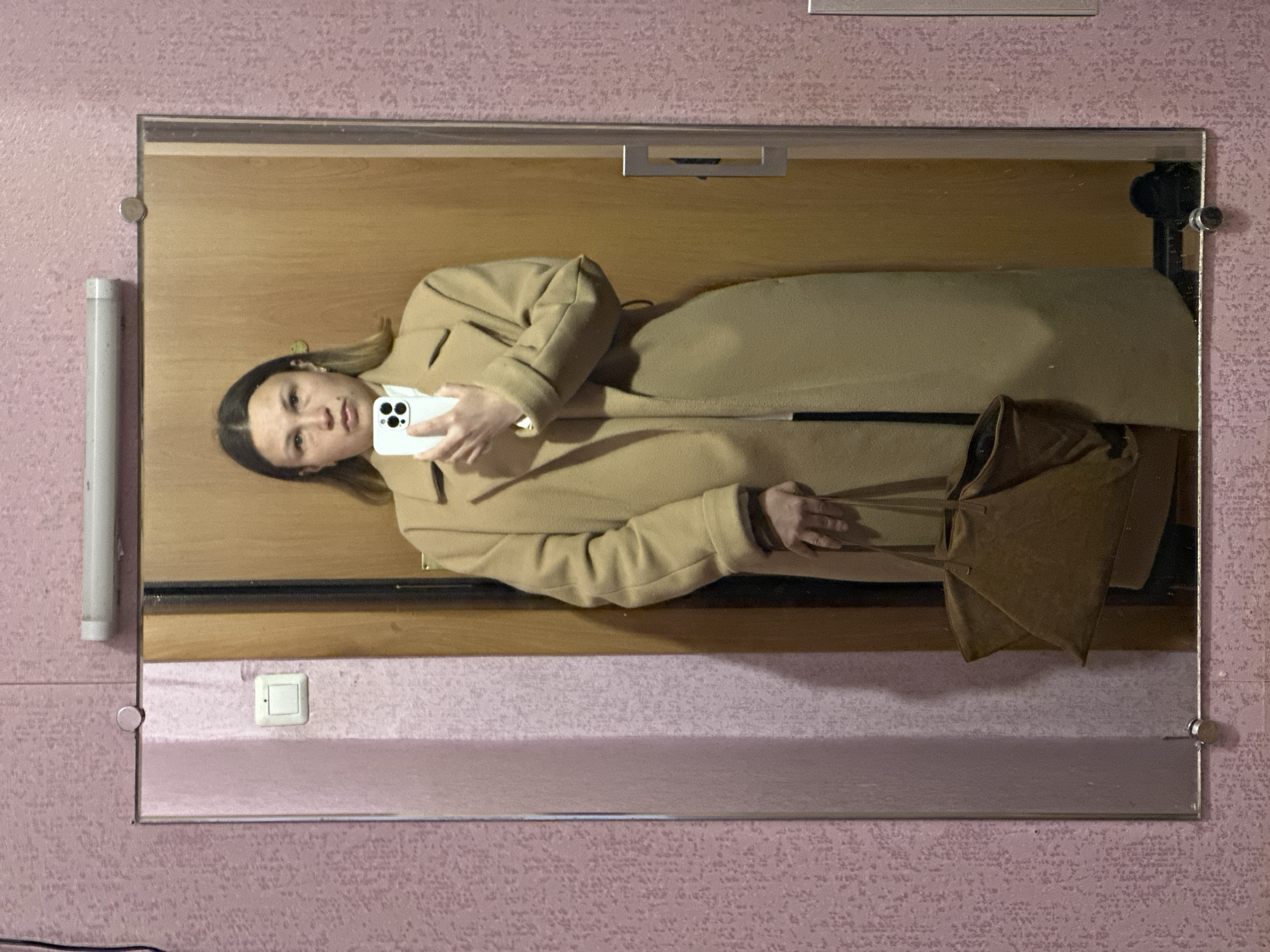 The image shows a woman in a beige overcoat holding a cell phone and a brown bag, reflected in a mirror. The mirror is mounted on a pink patterned wall with a light switch visible below the mirror. The location is Chaikovsky, Russia, as indicated by the light switch type used.