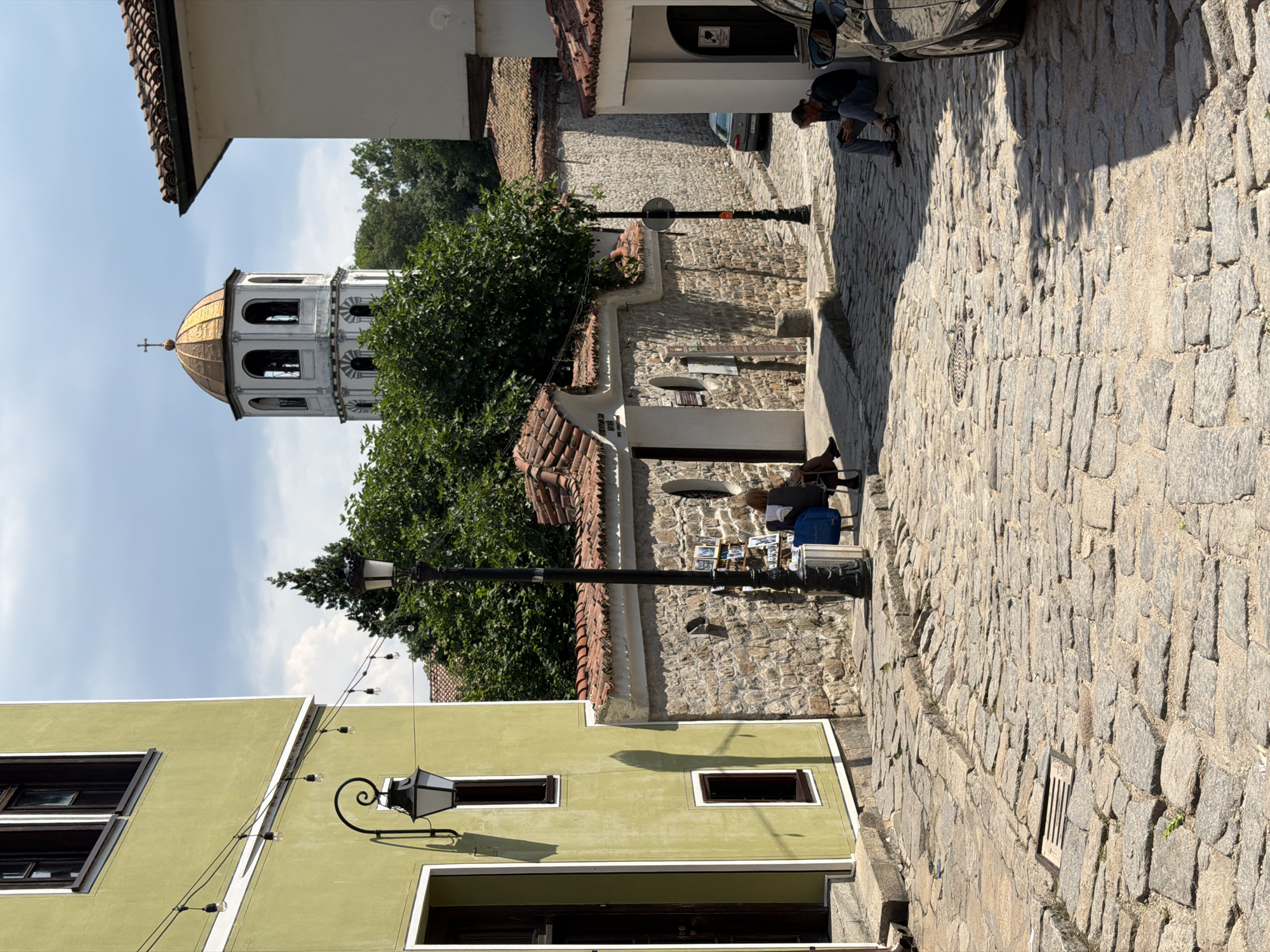 The image displays a daytime scene in the Old Town of Plovdiv, Bulgaria, characterized by historic architecture and a cobblestone street.
On the left, a tall, narrow building with a light green facade features two dark-framed windows and an open wooden doorway at ground level. A black, ornate wall-mounted lantern is affixed to its exterior. Above the lantern, a string of incandescent bulbs extends diagonally across the street.
The street, paved with irregular, light grey cobblestones, slopes gently downwards and curves slightly to the right. A rectangular metal drain grate is visible embedded in the cobblestones on the left side of the street.
In the midground, a stone wall with a reddish-brown tiled roof and white trim runs parallel to the street. This wall contains an arched entrance and a small circular window. A tall black lamppost stands in front of the wall, and a small rack displaying books is attached near its base. Adjacent to the lamppost, a person is seated on a low stool, engrossed in reading a book. A blue fabric bag rests on the ground beside them.
Further down the street on the right, another person is seated on a low ledge or the ground, looking down at a handheld device. A dark-colored vehicle is partially visible, parked on the far right. Beyond this, a silver car is parked further down the narrow, curving street.
In the background, rising above the tiled roofs and dense green foliage, is a prominent white bell tower. This tower features multiple arched openings and is capped with a golden, onion-shaped dome topped by a small cross. The sky above is partly cloudy blue.