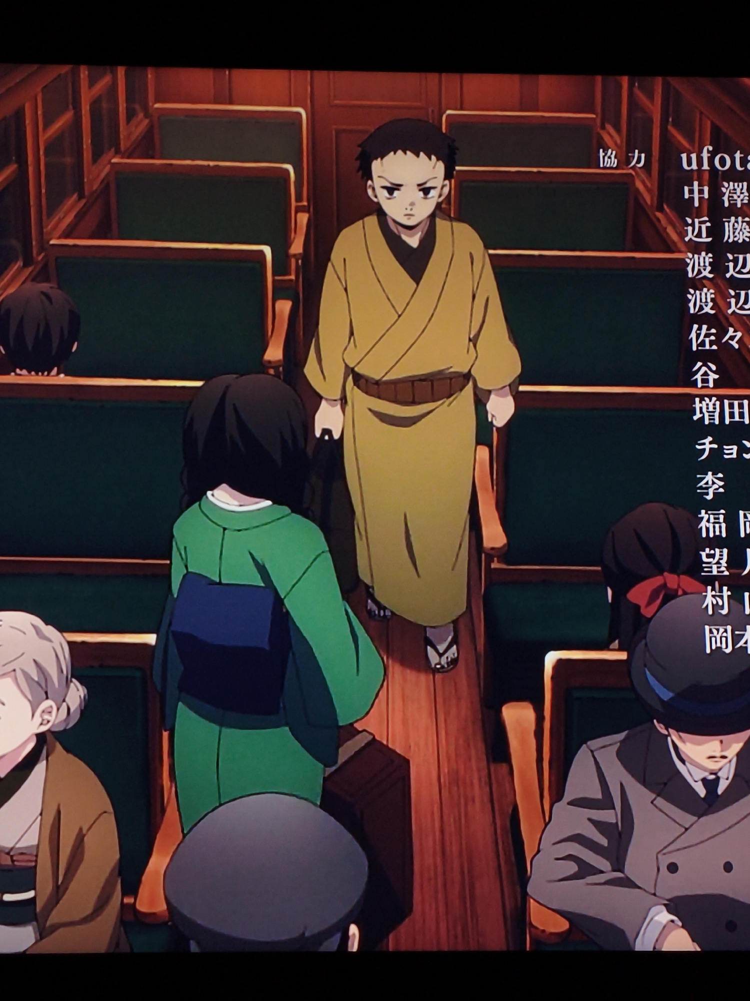 The image depicts an anime scene set inside a train carriage with rows of green seats. Several characters are visible, including a young boy in a yellow robe walking down the aisle, a girl in a green kimono, and a man in a grey suit wearing a hat. Japanese text, likely credits or subtitles, is displayed on the right side of the image. The environment suggests an indoor setting, possibly a historical or fantasy-themed train. There is no indication of any specific location within the city of Vladivostok, Russia.