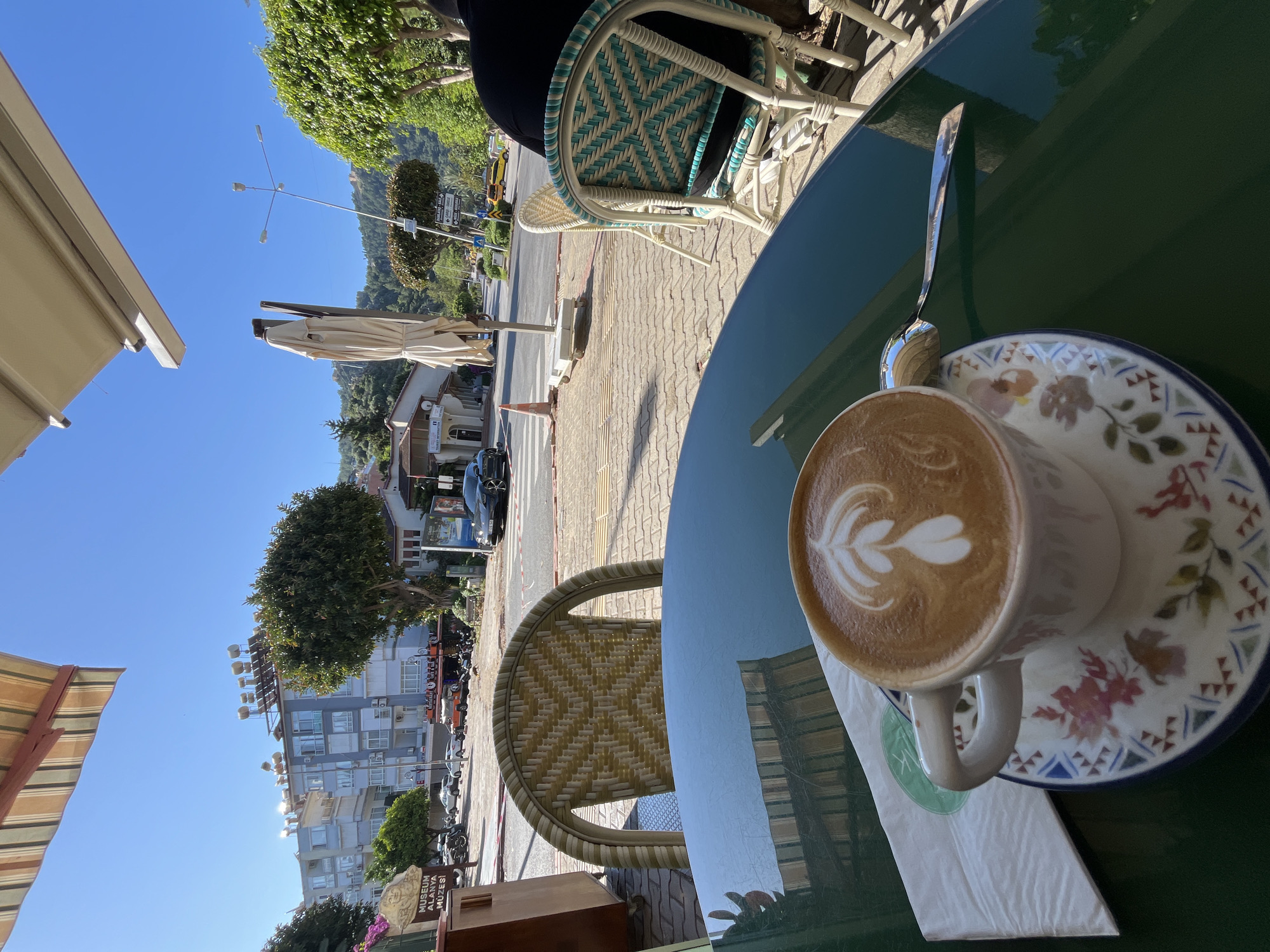 A coffee cup with latte art sits on a decorative saucer atop a green table on an outdoor patio. A spoon rests beside the cup. The table is next to woven chairs. Beyond the patio, the street shows buildings, parked motorbikes, cars, and a sign with "Museum Alanya Muzesi" visible. The location is in Alanya, Turkey. The weather is sunny with a clear blue sky.