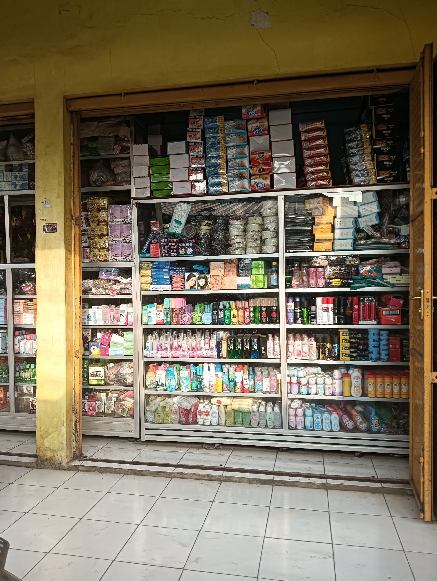 The image depicts a storefront in Tayu, Indonesia, displaying a variety of retail items. The store appears to be a small shop or convenience store, with merchandise organized on shelves behind glass doors and an open doorway on the right.
The storefront itself has a yellow, weathered exterior wall with visible cracks and peeling paint, suggesting age or exposure to the elements. The entrance has a wooden frame, painted yellow, with a yellow metal door on the right side, partially open, revealing more of the interior. The shop's interior is filled with shelves packed with products.
The merchandise includes:
* **Packaged goods**: Boxes of cookies, snacks, and other food items are stacked on various shelves. Examples include boxes with the brand name "GO!!" and stacked white and green boxes that appear to be food or household items.
* **Personal care and hygiene products**: Numerous bottles and containers of shampoo, conditioner, body wash, lotion, soap, and deodorant are prominently displayed. Many of these are in clear or colored plastic bottles. There are also several aerosol cans, likely deodorants or hair spray.
* **Household items**: Some shelves hold what appear to be cleaning supplies, detergents, or other household products in various packaging.
* **Cosmetics**: A few shelves feature cosmetic products, including small boxes and what look like facial creams or makeup. There are also images of women's faces on some packaging.
* **Miscellaneous items**: Other items, such as rolls of what might be toilet paper or paper towels, and some unidentifiable bagged items, are also present.
The lighting suggests it is daytime, with sunlight illuminating the exterior of the shop and casting shadows. The floor in front of the store is tiled with white square tiles. The overall impression is that of a busy, well-stocked local shop.