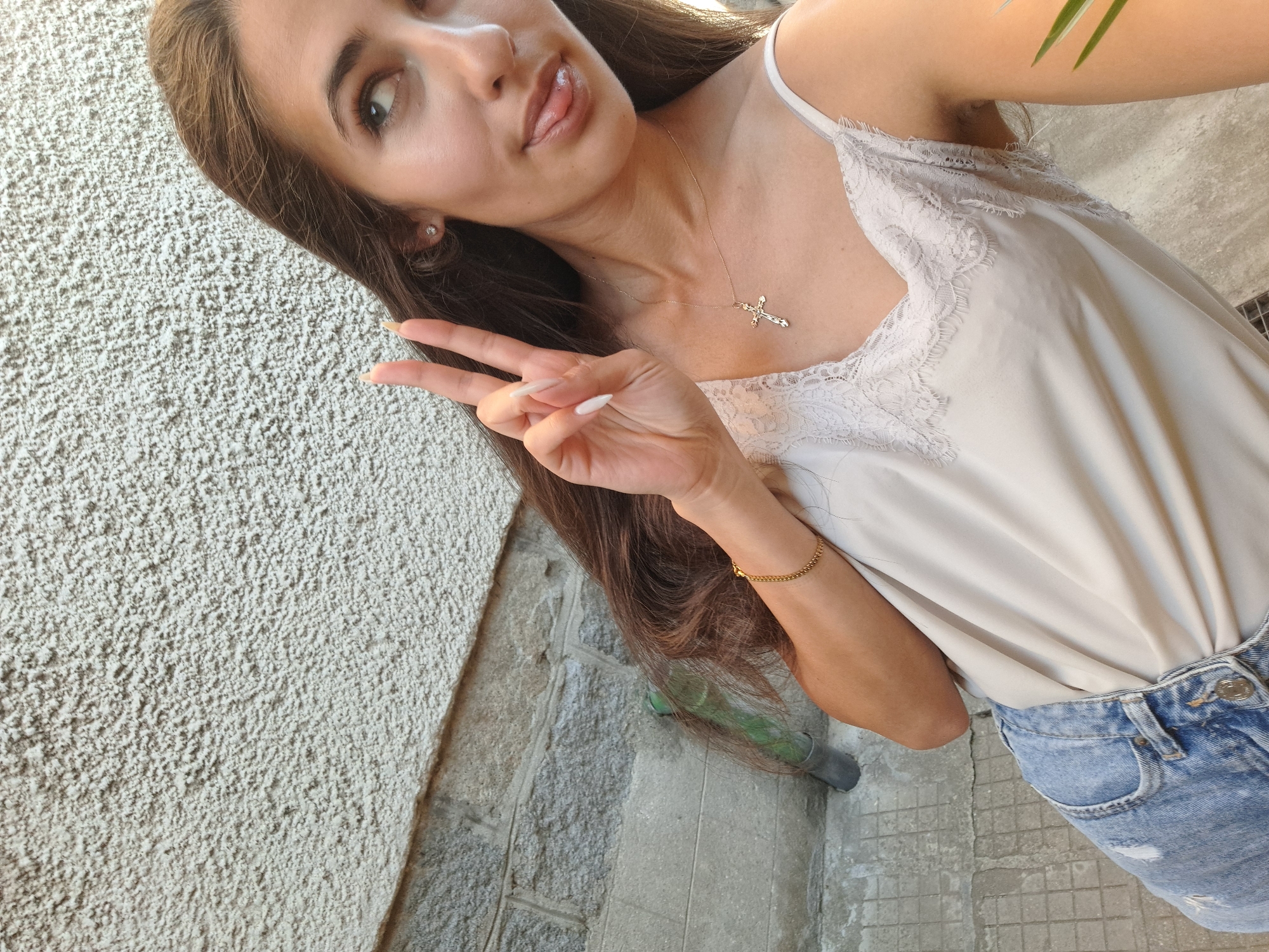 A young woman is taking a selfie outdoors, with a textured wall behind her and a stone wall to her left. She is wearing a light-colored camisole with lace trim and ripped denim jeans. She has long brown hair, and her left hand is raised, forming a peace sign. She is looking to the right and sticking her tongue out. She is wearing a delicate gold cross necklace and a gold bracelet. The environment appears to be a street or alleyway, with a drainpipe visible on the stone wall. The scene is well-lit, suggesting it is daytime.
