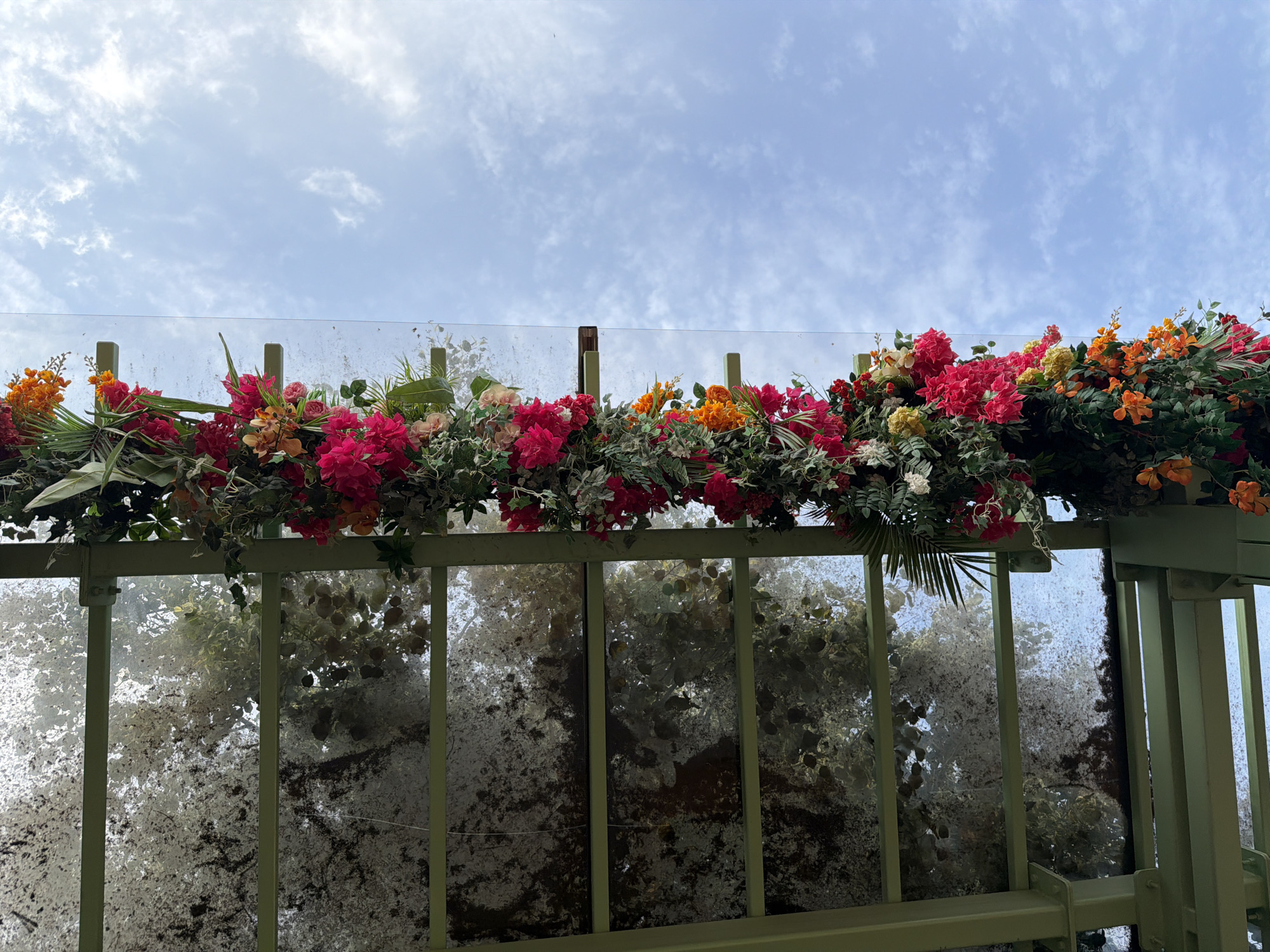 The image shows an overhead view of a glass structure with artificial flowers and foliage draped along its edge. The structure appears to be part of an outdoor setting, possibly a canopy or walkway, with a clear sky filled with scattered clouds visible above. The glass panels of the structure are somewhat obscured by what appears to be a layer of dirt or algae, and reflections of trees and foliage are visible within them. The artificial floral arrangement features a vibrant mix of pink, red, orange, and yellow flowers, interspersed with green leaves and palm fronds. The overall impression is one of decorative landscaping. There are no people or animals depicted in the image. The scene is not identifiable as being in Moscow, Russia, or any specific city.