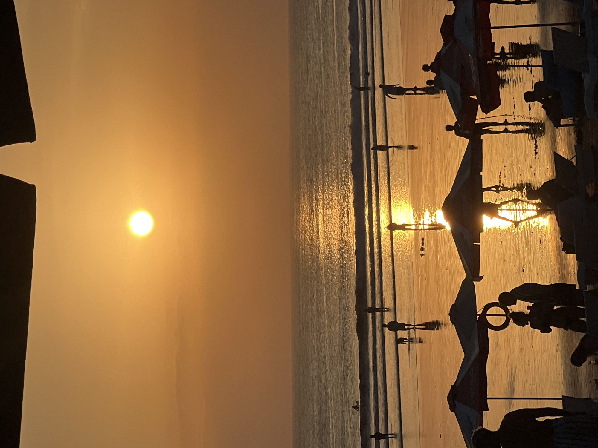 A wide shot captures a beach scene in Kuta, Indonesia, during sunset. The sun, a bright orb, is positioned in the upper center of the frame, casting a warm orange glow across the sky and reflecting on the wet sand and ocean. Silhouetted figures of people are scattered across the beach, some walking, others standing near beach umbrellas. In the foreground, a couple stands close together under an umbrella, while another person stands nearby. Further out, people are seen walking in the shallow water, and one person appears to be in the ocean. A dog is also visible on the sand. The ocean has gentle waves, and the water's surface shimmers with the sunset's reflection. Beach chairs and umbrellas dot the foreground, suggesting a popular tourist spot. The overall atmosphere is serene and picturesque, typical of a beach sunset.