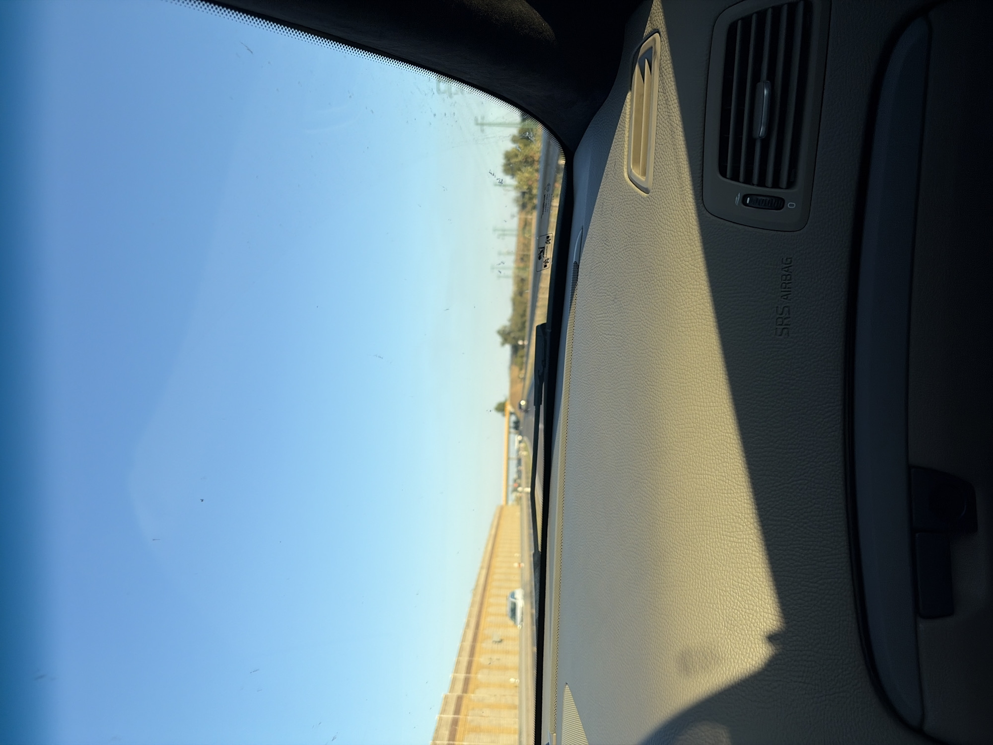 This image provides a view from the interior of a car driving on a highway, likely near Vlčkovce, Slovakia, given the context. The foreground is dominated by the textured dashboard of the car, with a prominent shadow cast across it from the left. To the right of the dashboard, an air vent and the "SRS AIRBAG" lettering are visible. The windshield shows some smudges and dust, through which the exterior scene is observed.

The exterior view reveals a clear, bright blue sky with a few faint specks. A highway stretches into the distance, flanked on the left by a tall, textured concrete wall or barrier, possibly a noise barrier, with vertical supports visible. Several cars are driving on the highway, appearing as blurry shapes due to motion. In the distance, the highway curves and an overpass is visible. Trees and vegetation line the sides of the highway further down. The lighting suggests it is daytime, possibly late afternoon, due to the long shadow on the dashboard.