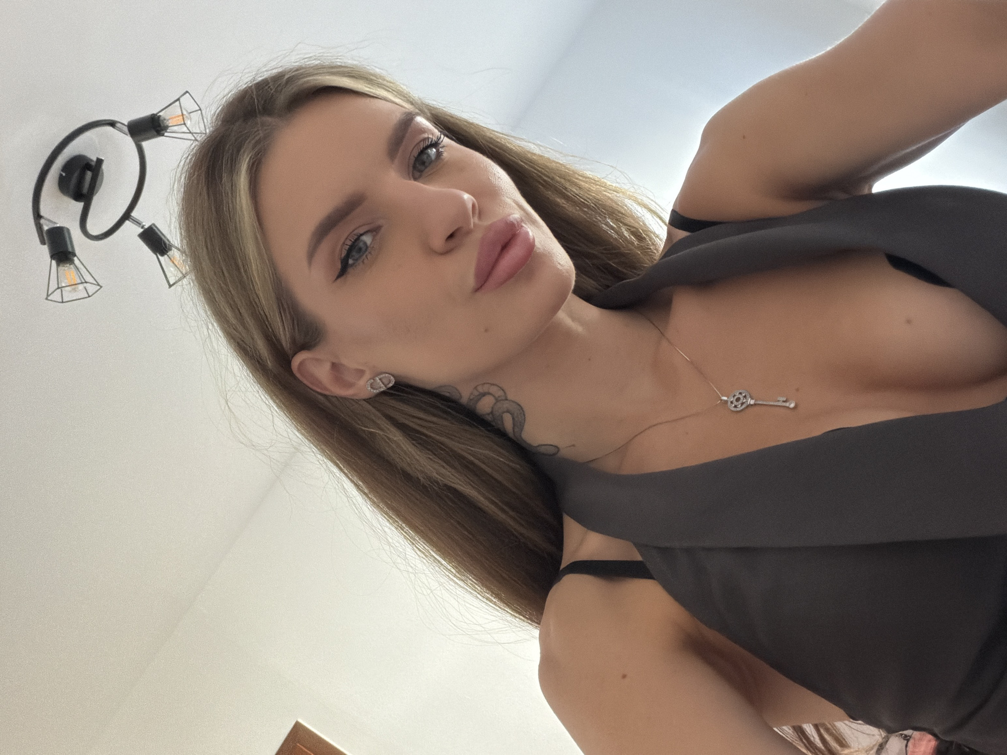 A woman with blonde hair and fair skin is looking into the camera with a slight pout. She has blue eyes and her eye makeup includes eyeliner. She is wearing a dark gray garment that reveals some cleavage and a silver key-shaped necklace. She has a tattoo on her neck and a small earring. In the background, there is a white wall with a modern-style light fixture with exposed bulbs. The location in the city of Berlin, Germany, cannot be determined.