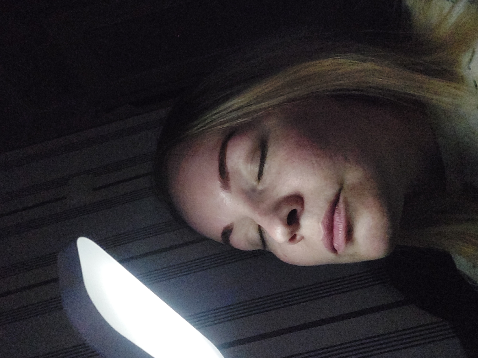 This media file features a close-up, slightly tilted view of a young woman's face, illuminated by a bright, white light source in the bottom left corner. The woman, with blonde or light brown hair, appears to be resting or sleeping, as her eyes are closed and her expression is peaceful. Her skin tone suggests she is fair-skinned, with visible cheeks and lips.
The setting is an indoor space, likely a bedroom, characterized by a dark background that suggests nighttime or a dimly lit room. Behind the woman, there is a dark surface with subtle, faint horizontal lines, possibly a wall, wallpaper, or a piece of furniture like a headboard. The bright light source is a lamp, positioned below and to the side of her face, casting an upward glow.
No specific activity is taking place other than the woman's apparent rest. There are no visible location cues within the image itself beyond indicating an indoor environment. No text is visible in the frame. The location context provided is Chaikovsky, Russia.