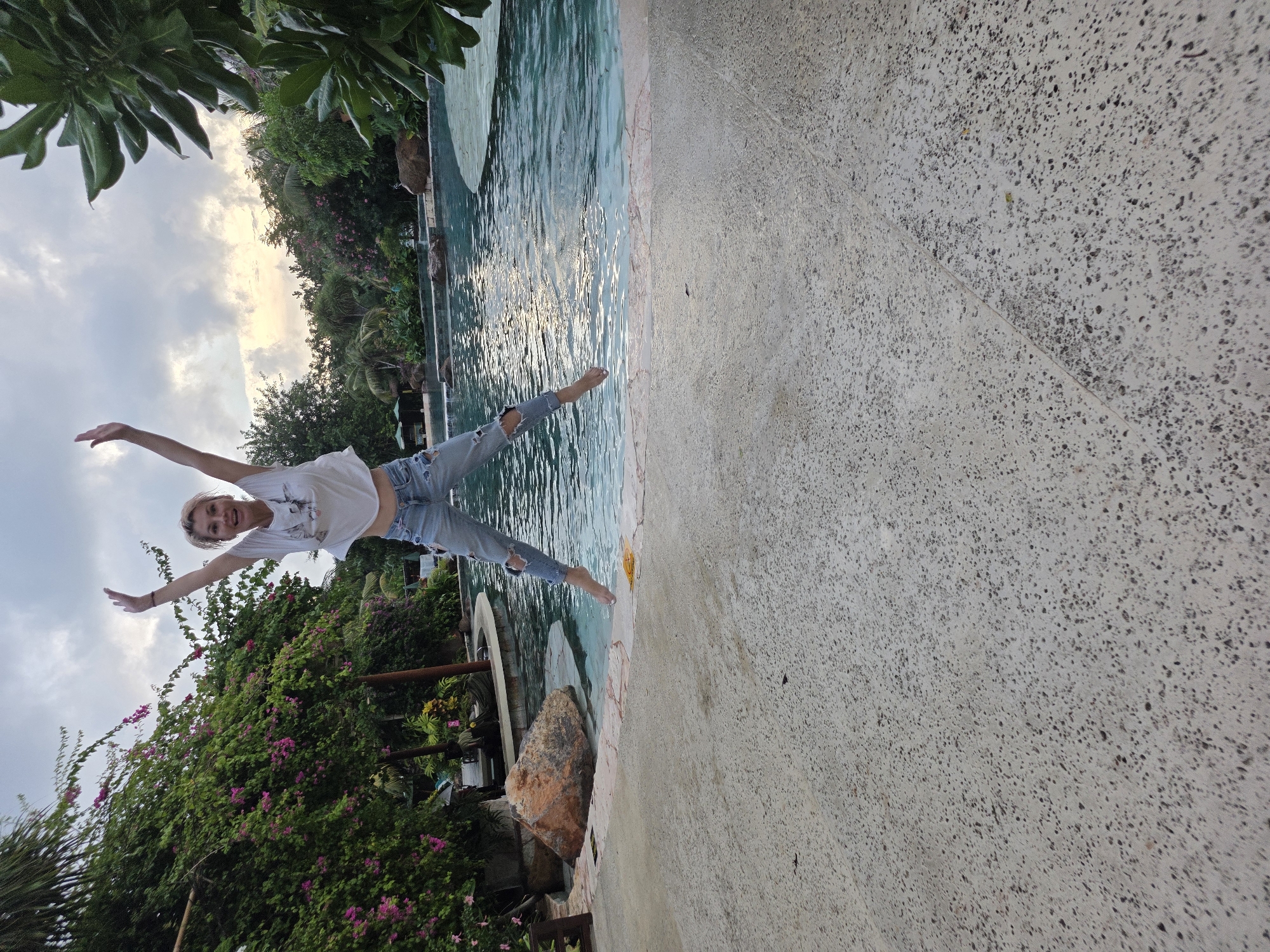 A woman is captured mid-air in a jumping pose next to a swimming pool. She is wearing a white t-shirt and ripped jeans, with her arms outstretched. The pool has a light blue hue with visible water ripples and is lined with pinkish stones. To the right of the pool is a concrete area. Behind the woman, lush greenery, including flowering plants and trees, creates a tropical backdrop. The sky is overcast, with a hint of light peeking through the clouds. The image is taken from an unusual angle, with the scene rotated. The location is potentially Kuta, Indonesia, given the vegetation and pool design.