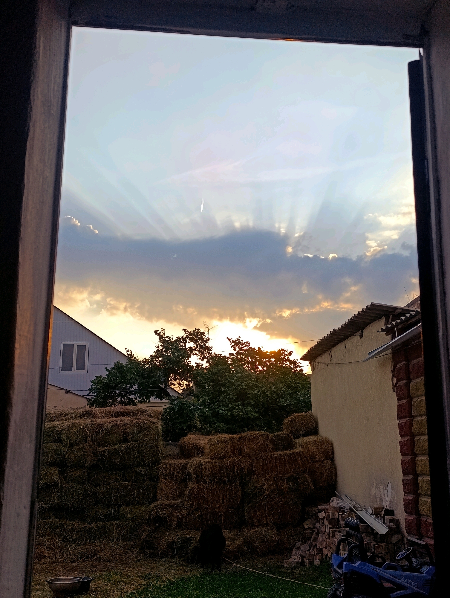 This is a photograph taken from inside a structure, looking outwards through an opening. The opening appears to be a window or doorway with dark wooden frames on the left and right edges, partially obscuring the view. The scene depicts an outdoor rural setting during what appears to be sunset.
In the foreground, on the left side, a large stack of hay bales is visible. These bales are rectangular and densely packed, forming a substantial pile. To the right of the hay bales, a dark-colored animal, possibly a cat, is sitting or lying on the grass. In front of the hay bales and slightly to the left, a dark, shallow bowl or basin is placed on the ground.
Further into the scene, a house with a light-colored exterior and a white window with two panes is visible on the left. Behind the hay bales and in the center of the frame, there are several trees with dense green foliage. The sun is setting behind these trees, casting a warm glow and creating visible sun rays emanating from behind the clouds.
On the right side of the image, a building with a light-colored wall and a corrugated roof is present. The wall has some visible imperfections and is bordered by a brick section on the right. Against the wall, a pile of bricks is stacked, and to the right of the bricks, a small blue toy vehicle, possibly an ATV, is parked.
The sky above is filled with clouds. Some clouds are dark and heavy, while others are illuminated by the setting sun with a golden hue. The sun's rays are visible, fanning out across the sky from behind the clouds. A faint streak, possibly a contrail from an aircraft, is visible in the upper right portion of the sky. The overall lighting suggests the late afternoon or early evening. The location is identified as Naryn City, Kyrgyzstan.