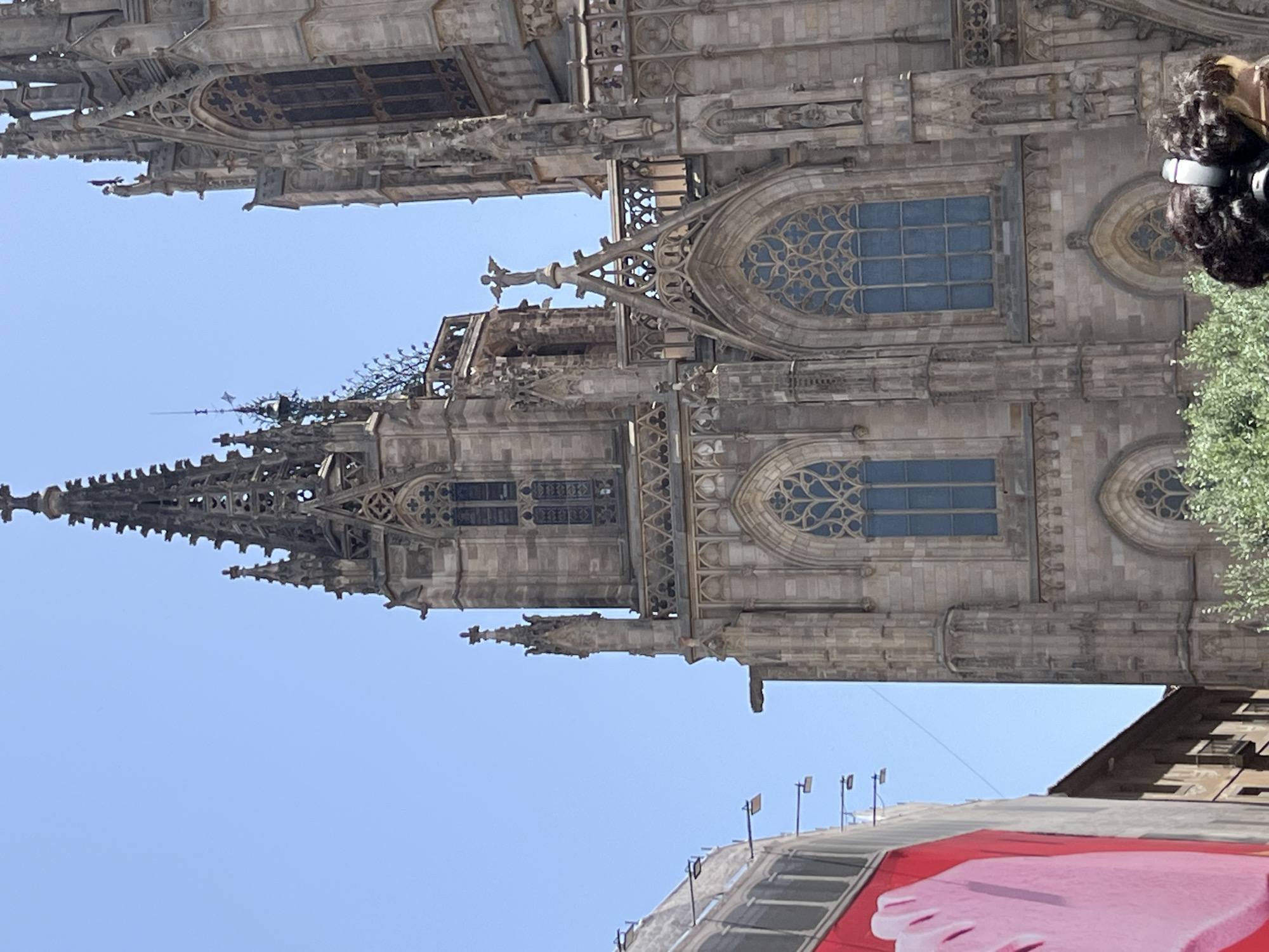 The media file is a photograph of the exterior of a large, ornate Gothic-style building, likely a cathedral, against a clear blue sky. The architecture is characterized by intricate stonework, spires, gargoyles, and pointed arches. Large windows with decorative tracery are visible, some filled with blue glass. The image is oriented vertically, suggesting it was taken with the camera held in portrait mode or that the building is being viewed from a low angle.
In the upper right corner of the frame, a person with dark, curly hair and headphones is partially visible, looking upwards and to the left. The person's hair is styled in an afro, and they are wearing a black top and headphones over their ears. A portion of a green tree is also visible in the foreground, to the right of the building.
Below the main structure, a partial view of another building with a sloped roof is visible, on which a large red banner with a pink graphic of a foot is displayed. Several light poles are also present on this lower structure. The bright, clear sky indicates it is daytime, likely sunny. The overall scene suggests an outdoor urban environment. No visible text is discernable in the photograph.