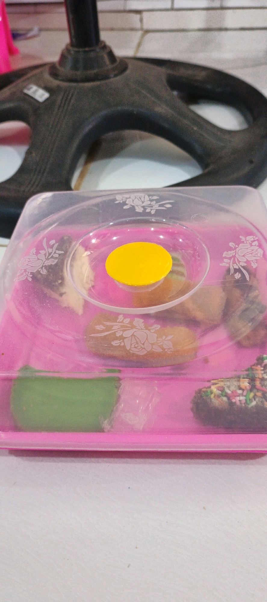 The image shows a close-up view of a clear plastic container with a pink base, filled with various small baked goods. A yellow circular sticker is placed on a raised clear plastic dome within the container. The baked goods appear to include at least one green-colored item, several brown items with colorful sprinkles, and other assorted baked goods. The container has embossed floral designs on its lid and base. In the background, the base of a black stand, possibly part of a fan or other appliance, is visible, and behind that, a white tiled wall. The scene is set on a white surface. The photograph does not contain any information to determine its location within the city of Simbur Naik, Indonesia.