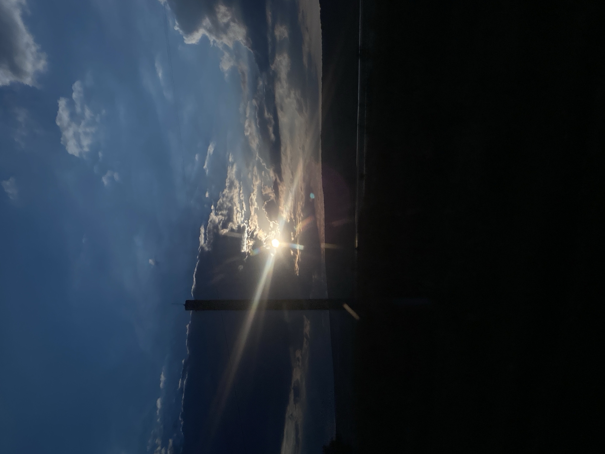 The image captures a dramatic sunset over a rural landscape. The sky is a deep blue, transitioning to lighter shades near the horizon, with scattered clouds that are illuminated by the setting sun. Beams of light emanate from behind a tall, slender object, possibly a utility pole or a small tower, which stands silhouetted against the bright sky. The sun itself is a brilliant, white orb peeking through gaps in the clouds, casting lens flare effects across the scene.
Below the sky, a dark, silhouetted band represents the horizon, which appears to be a gently rolling landscape or hills. In the very foreground, a completely dark area obscures any details of the immediate surroundings. There is a thin, horizontal line visible in the middle ground, possibly a body of water or a road, also rendered in shadow.
The overall atmosphere is serene yet powerful, characterized by the intense contrast between the bright sunbeams and the dark silhouettes. The specific location in Congaz, Moldova, is not visually identifiable from the image itself, but it depicts a natural scene under a dynamic sky, typical of a sunset in a rural setting. No people or specific actions are depicted.