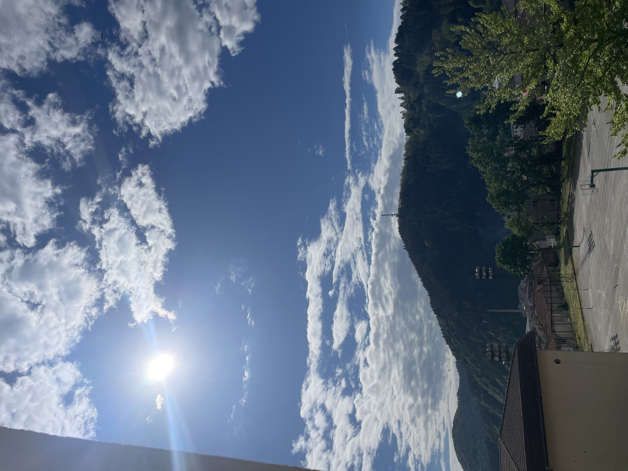 The image shows a view from an elevated position in Zlatograd, Bulgaria. The sky is predominantly blue with scattered cumulus clouds and a bright sun. A mountain covered in dense, green vegetation dominates the right side of the frame. The base of the mountain meets the edge of a building, possibly part of a school or sports facility, with a basketball court visible. The scene seems to be taken on a sunny day, with clear visibility.