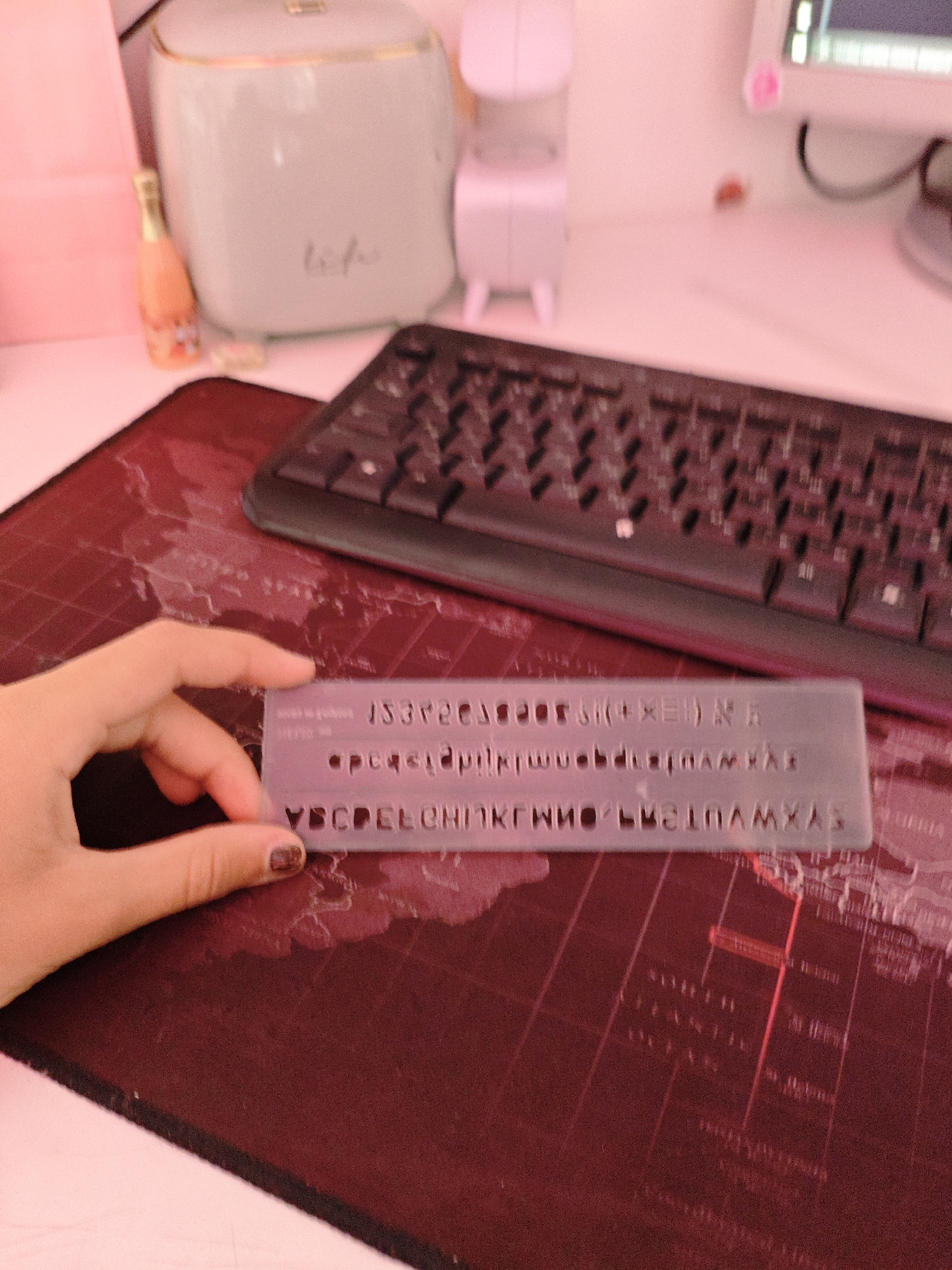 The image shows a close-up of a hand holding a transparent stencil with letters and numbers. The stencil is placed over a dark red surface that appears to be a map. In the background, a black keyboard is visible, along with a white cylindrical object and a small orange bottle. The lighting is predominantly pinkish, suggesting an artificial light source. There is no indication of the city of Трудовское сельское поселение, Ukraine in the image.