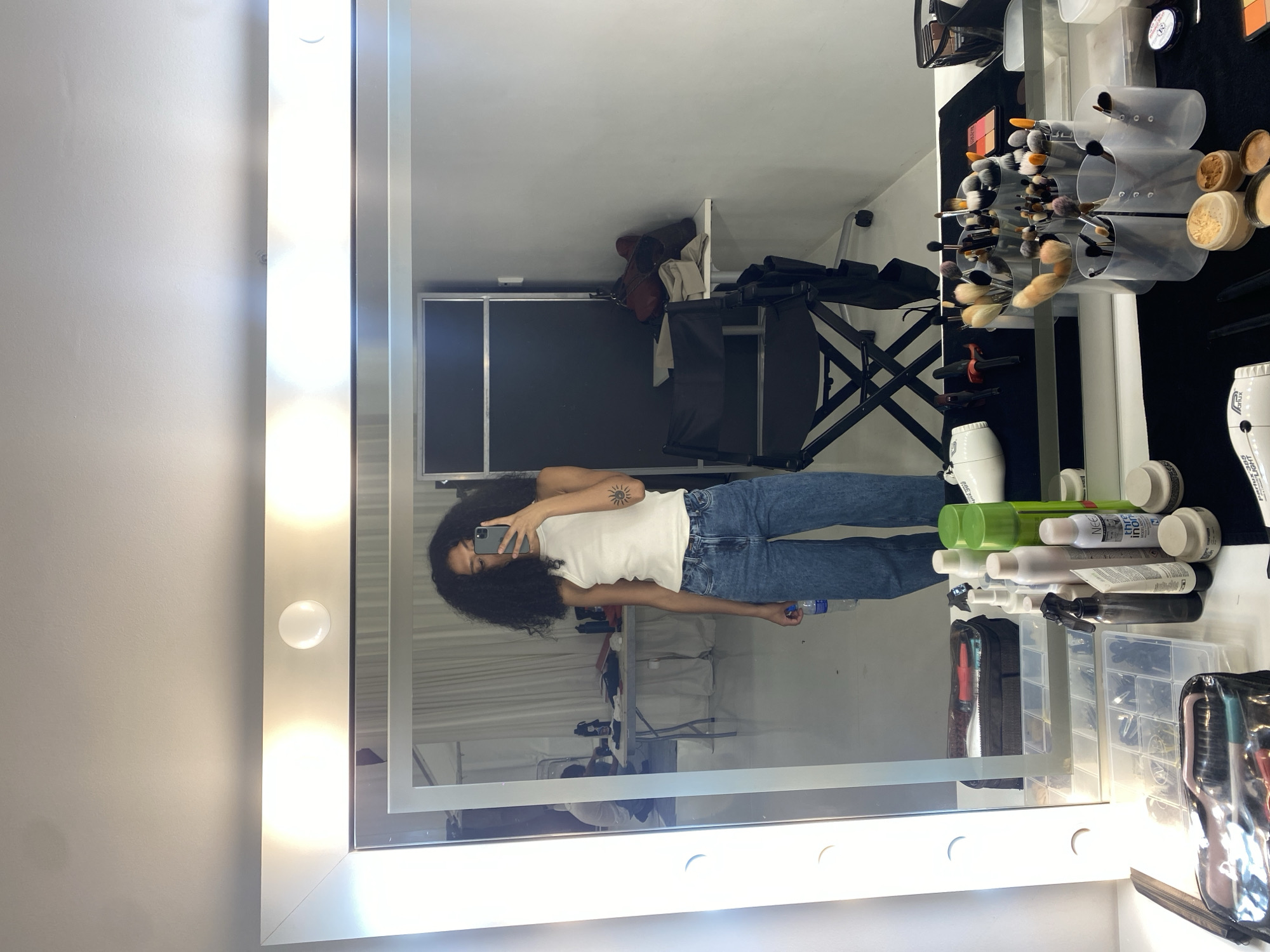 The image shows a person reflected in a makeup mirror with lights around the frame. The person is holding a smartphone and taking a picture of themselves. The person is wearing a white tank top and blue jeans. The mirror is surrounded by makeup brushes, bottles, and other beauty products. In the reflection of the mirror, there is a makeup chair and a backdrop. The location is likely a makeup studio or salon. There is no indication of the city of Porto Alegre, Brazil.