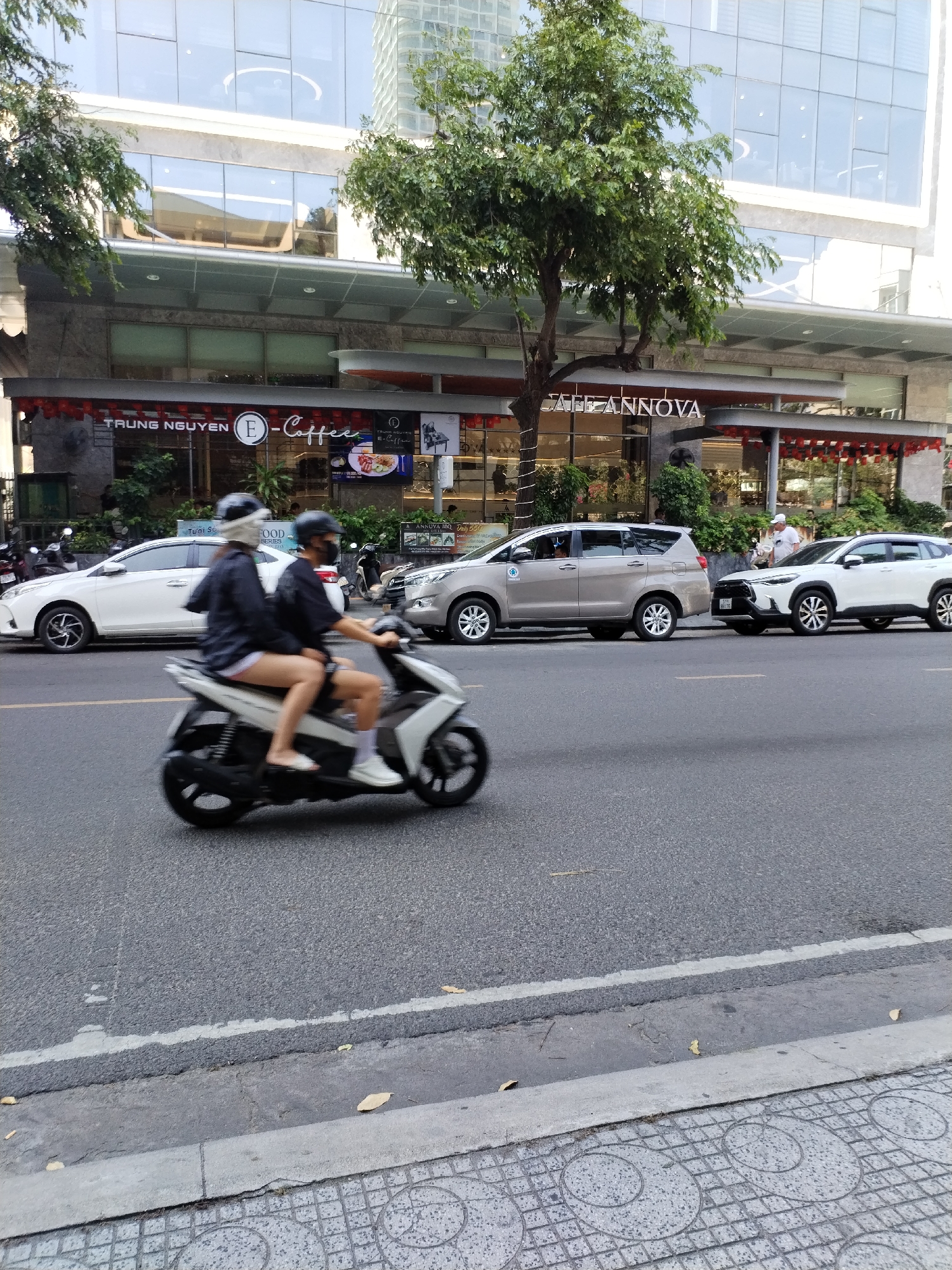 A daytime street scene in Nha Trang Ward, Vietnam, shows a couple riding a scooter in front of a building housing several businesses. The female passenger is wearing shorts, and both riders have helmets. Behind them, cars are parked along the street, and a pedestrian stands near the vehicles. The building has signs for "TRUNG NGUYEN E-Coffee" and "CAFFE ANNOVA". Trees line the street, and a modern building is visible in the background. The street is paved, and a tiled sidewalk is in the foreground.