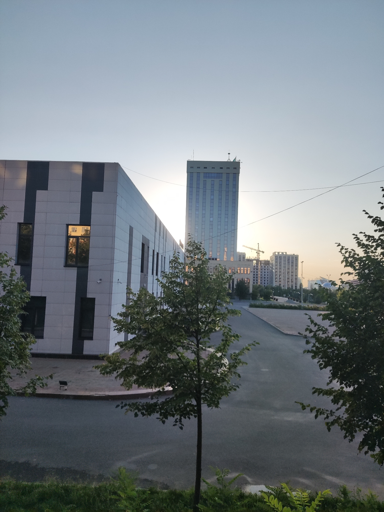 The image captures an urban scene in Shymkent, Kazakhstan, featuring modern architecture and a clear sky. In the foreground, a low-rise building with a white facade accented by dark vertical stripes is visible on the left. Its windows reflect the surrounding environment and the sky. To the right, a paved area, possibly a road or parking lot, leads towards a backdrop of taller buildings.
Dominating the mid-ground is a tall, slender skyscraper with a blue-tinted glass facade, characterized by numerous rectangular windows. The skyscraper appears to be illuminated from within, suggesting daytime activity. Several other multi-story buildings are scattered across the cityscape, some appearing to be under construction, indicated by the presence of a crane. Trees with green foliage are strategically placed, framing the scene and adding natural elements to the urban landscape. The sky is a pale blue, transitioning to a lighter hue near the horizon, suggesting it might be early morning or late afternoon. The overall impression is one of contemporary urban development.