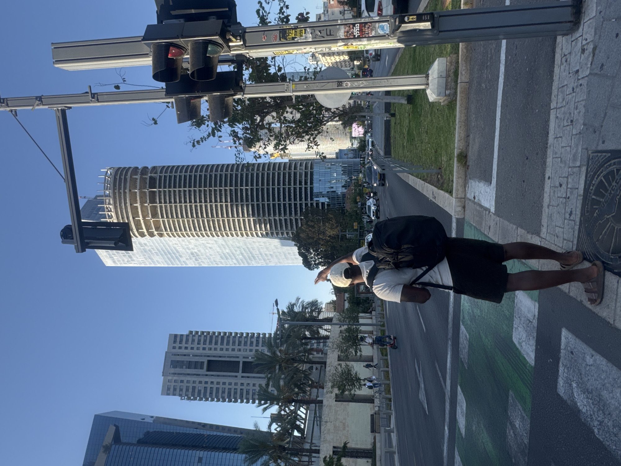 The image captures an urban street scene in Tel Aviv, Israel, on a clear, sunny day. The main subject is a person, appearing male, with dark skin, wearing a white t-shirt, black shorts, a black backpack, and sandals. They are also wearing a light-colored cap or hat and have their right arm raised, seemingly pointing towards something in the distance or gesturing.
The person stands on a sidewalk, next to a street featuring white markings and green bicycle lane indicators. A prominent manhole cover with circular patterns is visible on the sidewalk to their right. In the immediate foreground, a traffic light pole stands tall, holding multiple traffic light units, one of which shows a red light. The pole is adorned with various stickers, some featuring Hebrew text, including what appears to be "ישראל" (Israel) and possibly "תל אביב" (Tel Aviv), along with numbers like "084" and "18".
In the background, a modern cityscape unfolds under a bright blue sky. Several high-rise buildings dominate the skyline, including a distinctive curved, ribbed skyscraper (likely the Azrieli Sarona Tower) and other glass-fronted structures. Lush greenery, including palm trees, lines parts of the street and separates the pedestrian areas from the road. Cars and other vehicles are visible on the road further down. The overall impression is that of a bustling, contemporary city environment.
