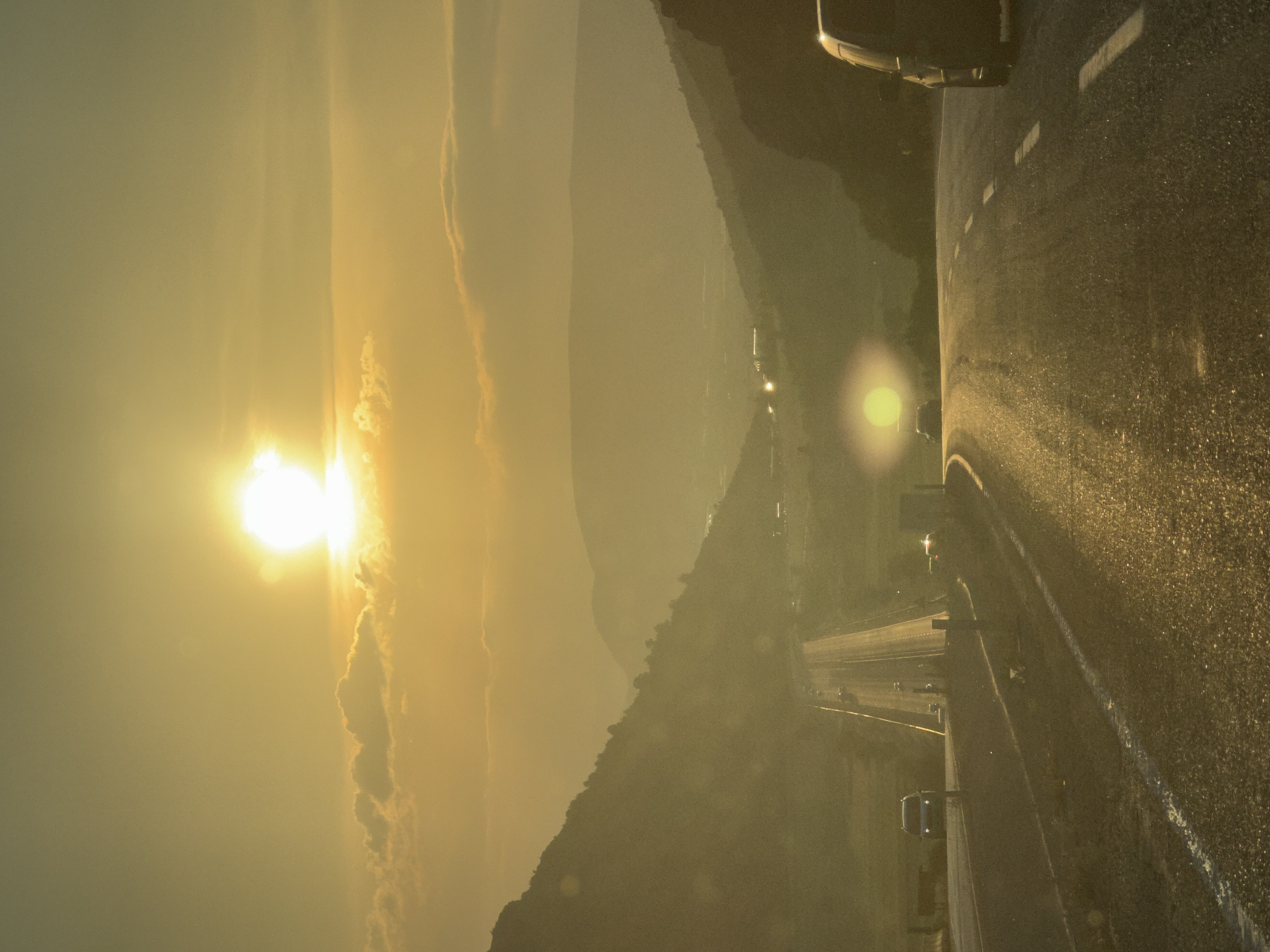 This detailed image displays a highway traversing a mountainous valley in Korkuteli, Turkey, during the golden hour, characterized by a dominant warm, golden-yellow illumination. A highly luminous sun, accompanied by significant optical flare, is positioned low in the upper-left quadrant of the sky. Scattered cumulus clouds are visible around the sun and along the horizon.
The landscape features a steep, vegetated mountain slope on the left, extending from the mid-ground towards the upper-left. Multiple layers of distant mountains are visible, exhibiting atmospheric haze that diminishes their detail and intensity towards the horizon. A valley floor is discernible between these geographical features, containing faint indications of human structures or settlements.
The foreground and mid-ground are dominated by an asphalt road. A straight segment of this road, likely a multi-lane highway, extends from the mid-ground on the left into the distant valley. Several vehicles are present on this segment: a light-colored sedan or van travels in the left lane towards the viewer, and multiple other indistinct vehicles are visible further down the road, some with discernible headlights or taillights. A separate segment of the road curves into the immediate foreground on the right, where the rear portion of a dark-colored vehicle (possibly an SUV or van) is partially visible on the far right edge of the frame.
The road surface is highly reflective, showing specular highlights from the low sun, which also casts a distinct, circular secondary flare onto a distant part of the road within the valley. No individuals are explicitly visible. The overall atmospheric condition includes significant haze, contributing to the diffused light and softened appearance of distant elements.