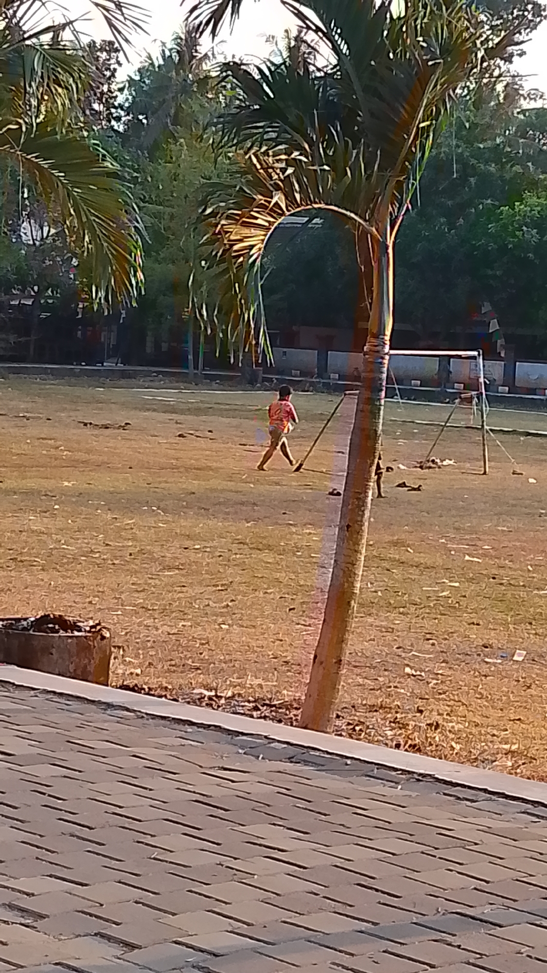 In Karanglangu, Indonesia, this image depicts an outdoor scene with a dry, grassy field, likely a sports ground, in the foreground. A young person in a red shirt and light-colored shorts is visible on the field, appearing to be in motion, possibly playing or practicing. To the right, a crude soccer goal with wooden posts is set up. A tall, slender palm tree stands prominently in the mid-ground, its fronds reaching towards the sky. The background shows a dense line of trees and some structures, suggesting a park or recreational area. The ground in the immediate foreground is paved with interlocking bricks. The lighting indicates it's likely late afternoon or early morning, with warm sunlight casting shadows.