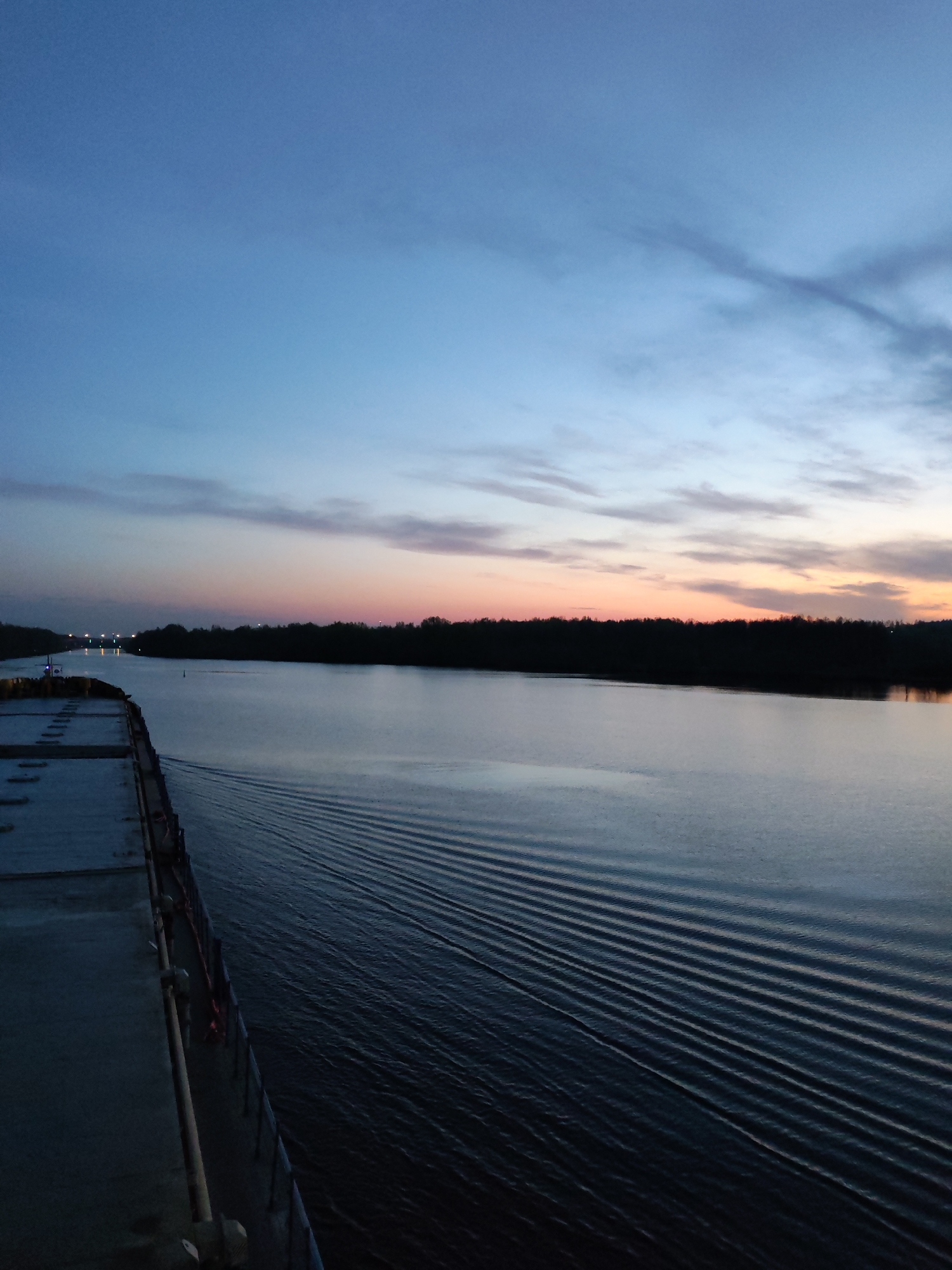 The image shows a river scene at dusk or dawn. The sky is a gradient of blue and pink hues, indicating a transition between day and night. A dark strip of trees lines the horizon on the opposite bank of the river. The river's surface is calm, with visible wake patterns from a moving vessel, which is partially visible on the left side of the frame. The vessel appears to be a large ship, possibly a barge. Lights are visible in the distance, suggesting a city or industrial area along the river. The location is likely on the Mississippi River, within the region of Vacherie, Louisiana.