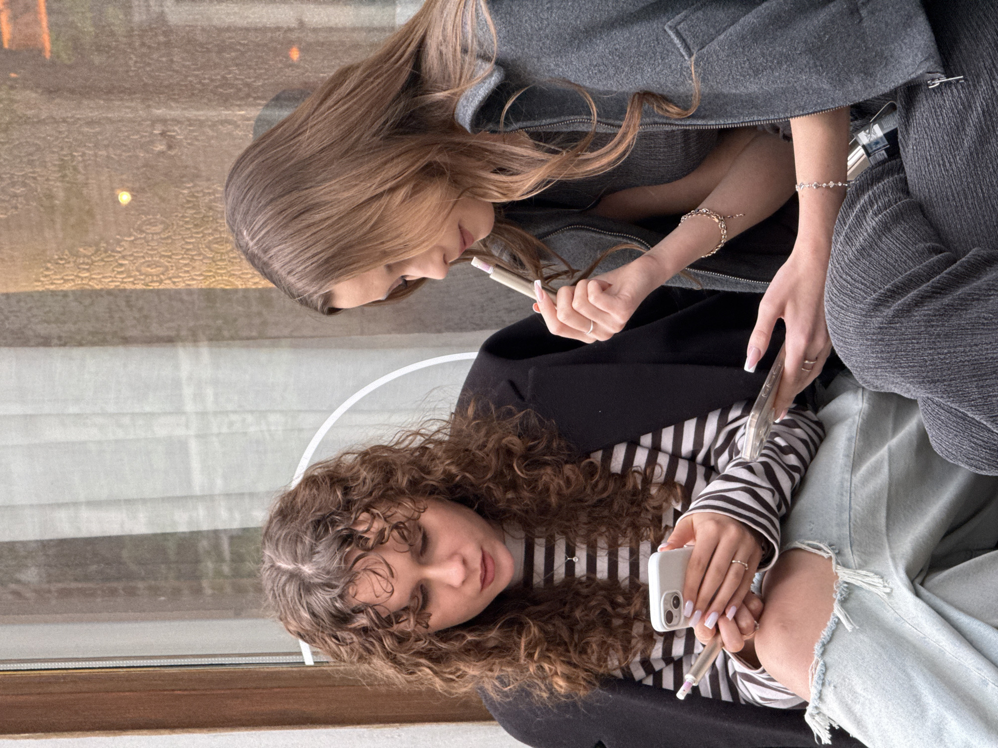 Two women are seated indoors. The woman at the top of the image has long brown hair and is wearing a gray jacket. She holds a pen or cosmetic item in her hand. The woman at the bottom of the image has curly brown hair, is wearing a black and white striped shirt, a black blazer, and ripped jeans. She is holding a white smartphone. The background shows a window with condensation on the glass. It's unclear where in Belgrade, Serbia the photo was taken based on the image alone.