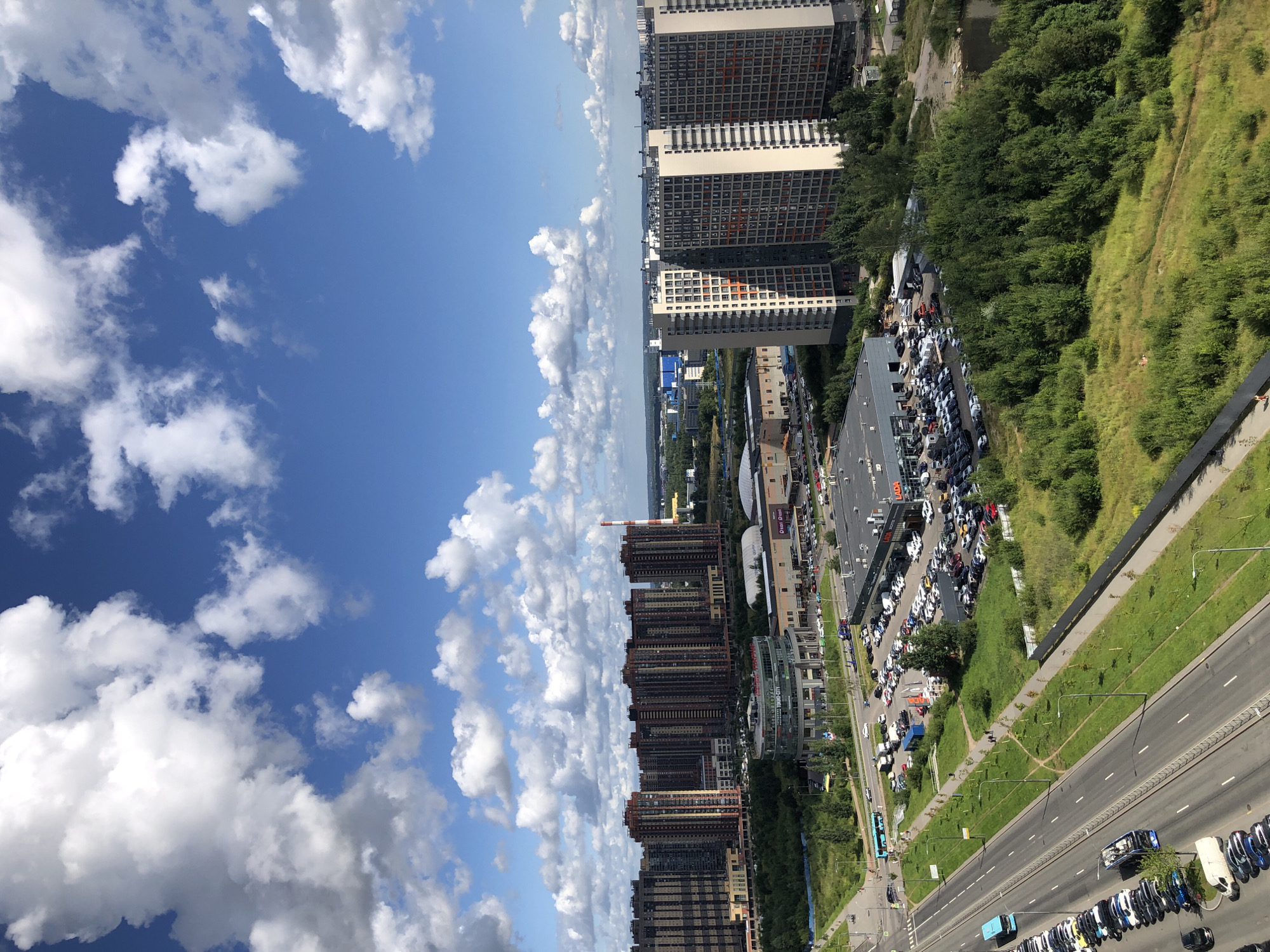 This is a high-angle, full shot of a suburban city landscape under a partly cloudy blue sky. In the foreground, a multi-lane road runs diagonally across the frame, with cars and buses driving and parked along its sides. To the right of the road, a large, flat-roofed commercial building, possibly a car dealership, is visible, with a vast parking lot filled with vehicles in front of it. Beyond this, a dense area of green trees and vegetation occupies the midground. Further back, several multi-story residential buildings and a cylindrical, glass-fronted structure are prominent. In the distance, more industrial or commercial buildings and a red and white striped chimney can be seen, blending into the hazy horizon. The overall scene depicts a mix of urban development and green spaces.