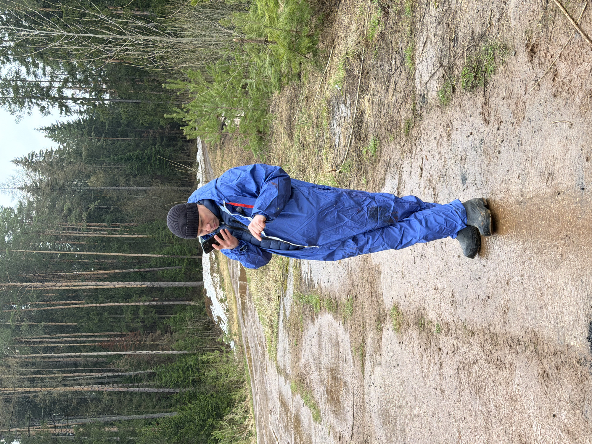 A person in a blue jumpsuit and black beanie is standing on a dirt path in a wooded area. The person is looking at a smartphone. The path is muddy and there is some snow on the ground. The trees are tall and mostly bare. The person is located in Прикамский, Russia.