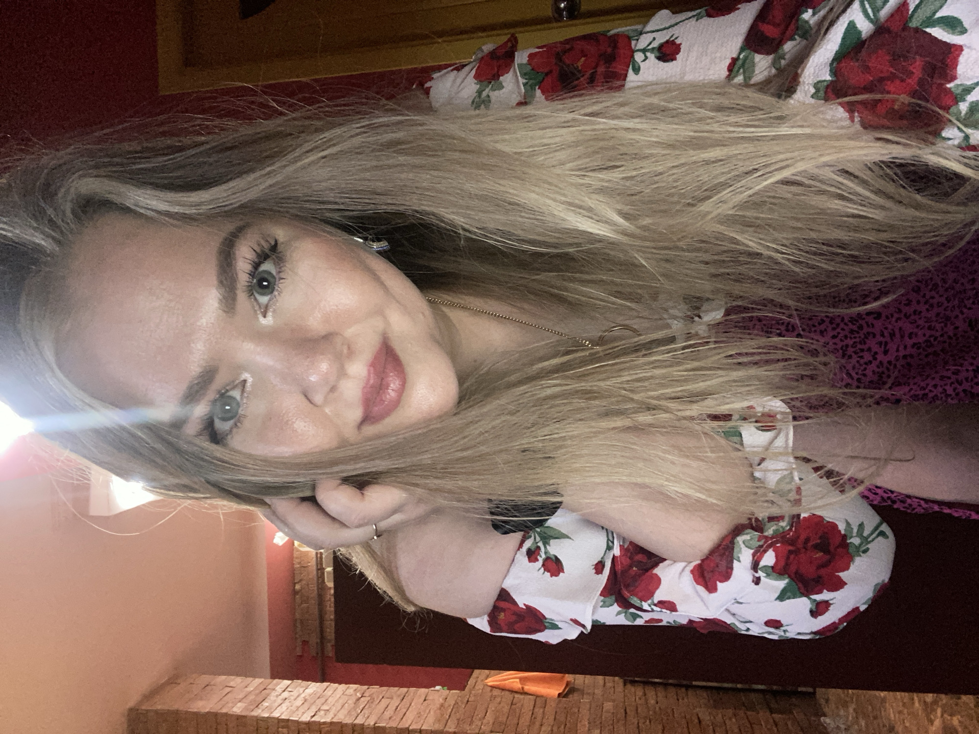 The image shows a young woman with blonde hair and blue eyes, wearing a floral-print off-the-shoulder top and a pink patterned dress. She has makeup on, including lipstick and eye makeup, and is wearing a necklace and earrings. Her hand is resting on her head, and she is looking at the camera. The background appears to be an indoor setting, possibly a room in a house or apartment, with a brick wall visible on the left and a red wall and a doorway on the right. The lighting suggests it is indoors. The image does not provide any visual information to determine a location in the city of Yerevan, Armenia.