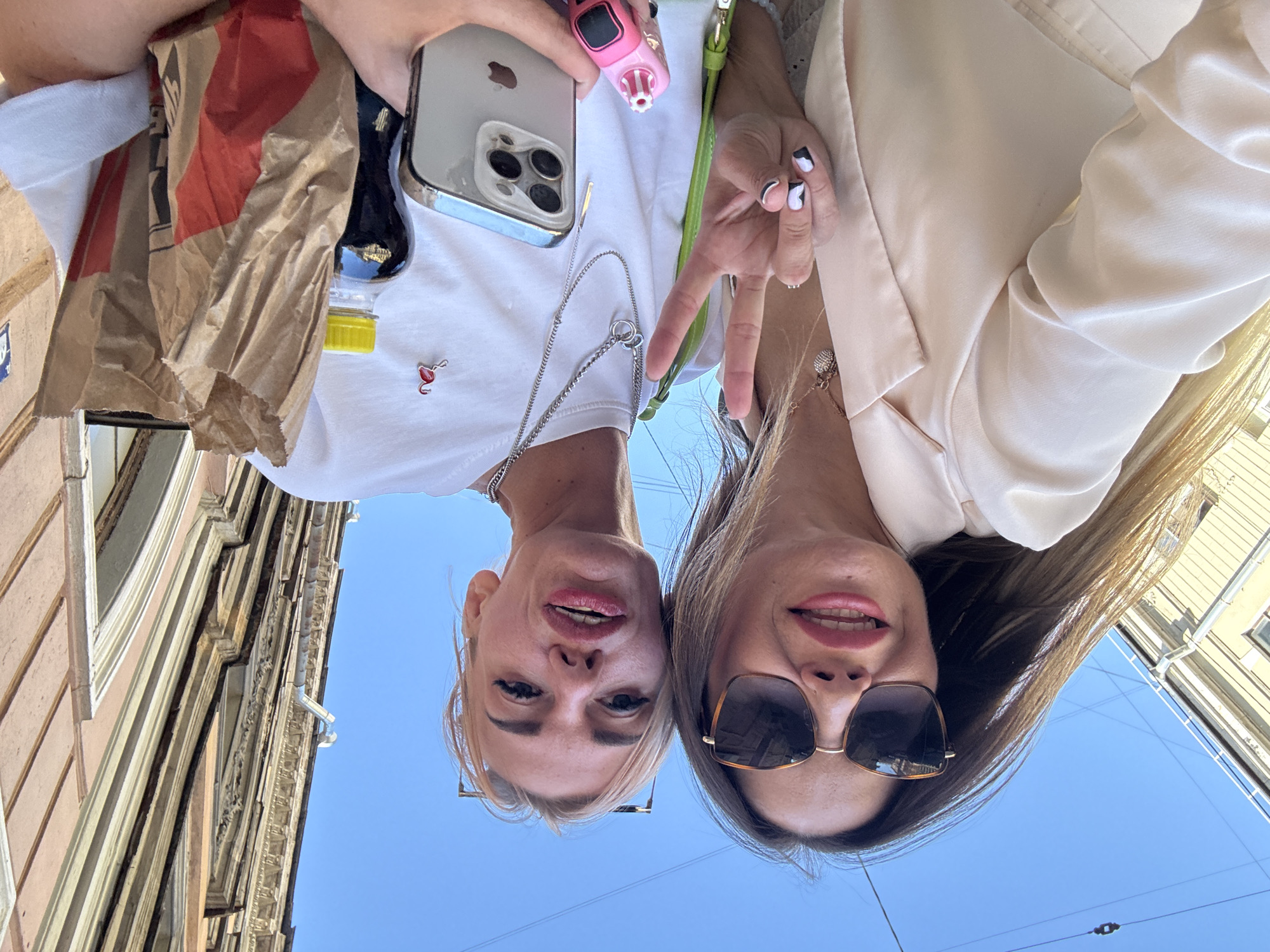 Two women are taking a selfie outdoors on a sunny day in Saint Petersburg, Russia. The woman on the left, wearing sunglasses and a cream-colored blazer, holds her hand up in a peace sign. The woman on the right, wearing a white t-shirt, holds a mobile phone and a paper bag containing food. A bottle of dark liquid with a yellow cap is also visible. The background shows the blue sky and the side of an old building with ornate architectural details. The image is taken from a low angle, looking up towards the sky.