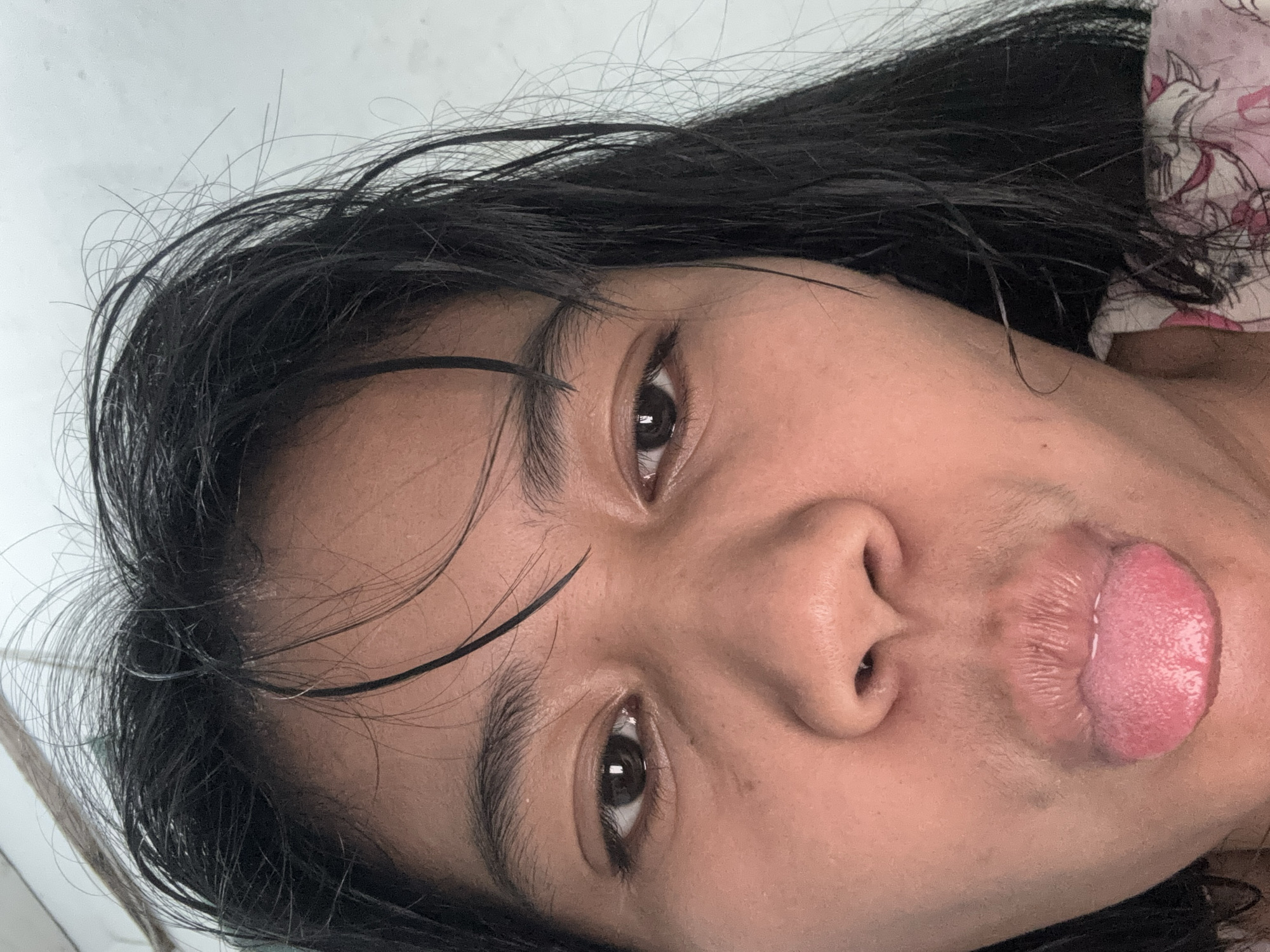 A close-up selfie of a woman's face is shown. She has dark hair that is wet and unkempt, framing her face. Her eyes are open and looking towards the camera. Her mouth is open, with her tongue sticking out. She appears to be indoors, with a plain white wall visible in the background. The image is likely from North Jakarta, Indonesia, given the presence of a person of Indonesian descent and the general environment. The lighting suggests it was taken during the day.