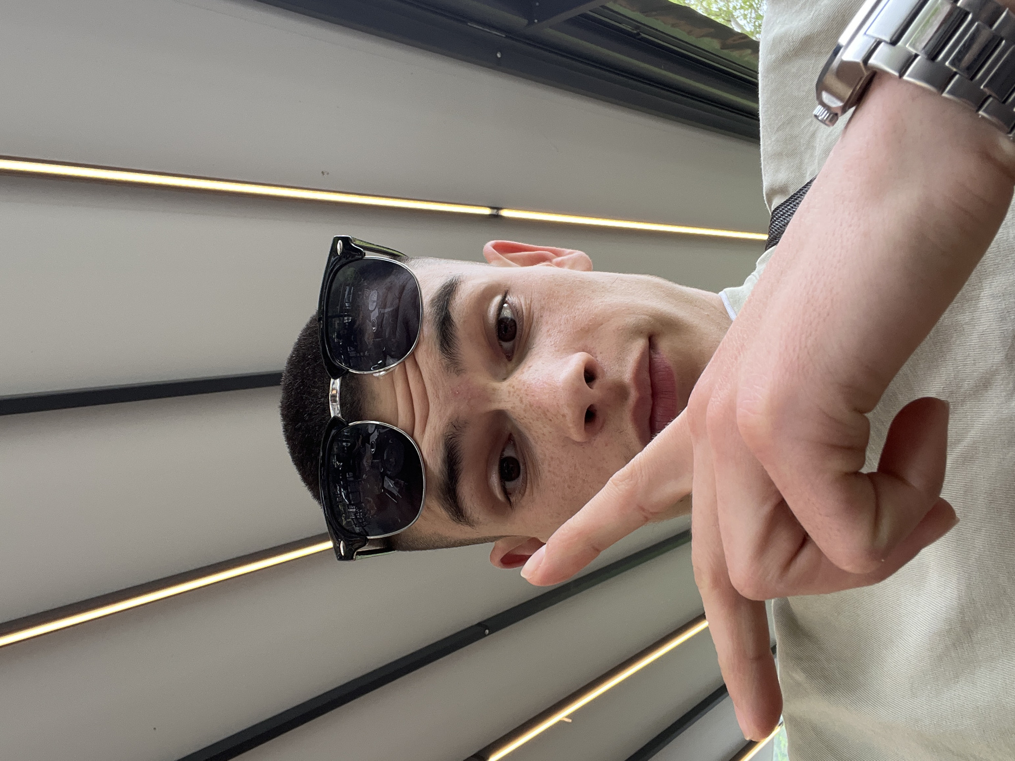 A low-angle shot shows a person with short, dark hair wearing sunglasses. The person has light skin and freckles. Part of their face, including their nose and one eye, is visible. The person's right hand is extended toward the camera with the index finger pointing downwards. The wrist of the same arm is adorned with a silver watch.
The background consists of a roof structure with parallel black beams and recessed linear lights running along the beams.
I cannot make a determination regarding Bachkovo, Bulgaria from the provided image and lack of context.