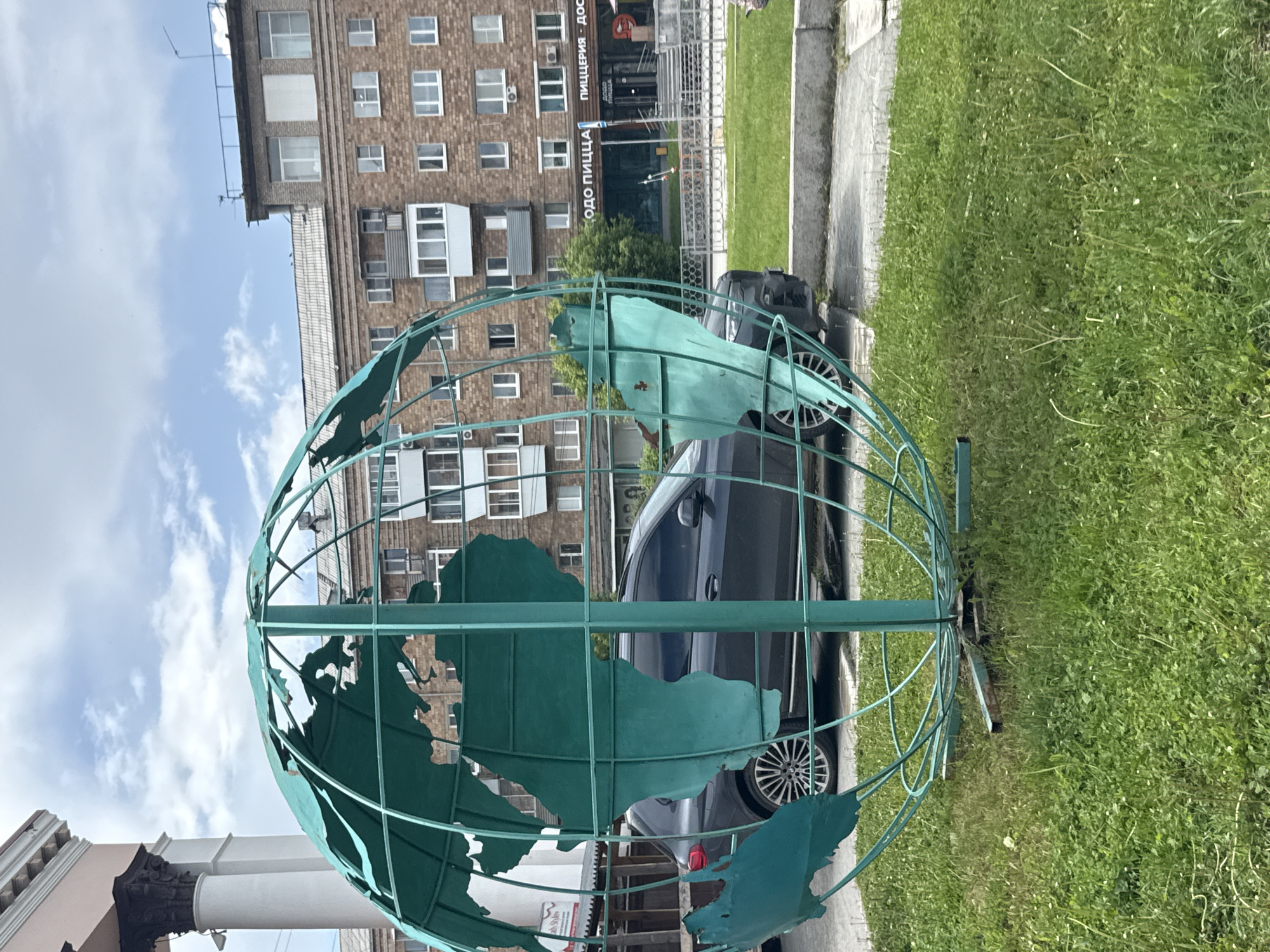 The image depicts an outdoor urban scene in Yekaterinburg, Russia, specifically on Lenin Avenue (Prospekt Lenina) near building number 51.
In the immediate foreground, a large, free-standing metal sculpture of a globe is prominent, positioned on a patch of vibrant green grass intermingled with small white flowers. The globe, approximately 2-3 meters in diameter, is constructed from teal-colored metal, forming a spherical framework with cut-out shapes representing continents, notably Africa and South America. The paint shows signs of weathering, with some peeling and rust spots visible. A thick vertical pole supports the globe from its center, anchored to a rectangular metal base on the ground. A concrete curb separates the grassy area from a paved surface on the right.
In the midground, directly behind the globe, a dark-colored SUV, possibly a BMW, is parked, with its side and rear wheels visible through the globe's structure. To the far left, a portion of a classical-style building with white fluted columns, a dark ornate capital, and a light-colored frieze is partially visible. Further behind the vehicle and to the right, a multi-story brick building with reddish-brown facade and numerous uniformly sized windows and some enclosed balconies dominates the mid to background. This building exhibits typical Soviet-era residential or mixed-use architecture.
In the background, at ground level of the brick building, a commercial storefront is visible with large glass windows. Cyrillic text on the signage reads "ОДО ПИЦЦА" and "ПИЦЦЕРИЯ - ДОС", clearly indicating a "Dodo Pizza" establishment. The brick building extends upwards, culminating in a flat roof equipped with various antennas and other structures against a partly cloudy, light blue sky. No people are visible in the frame.