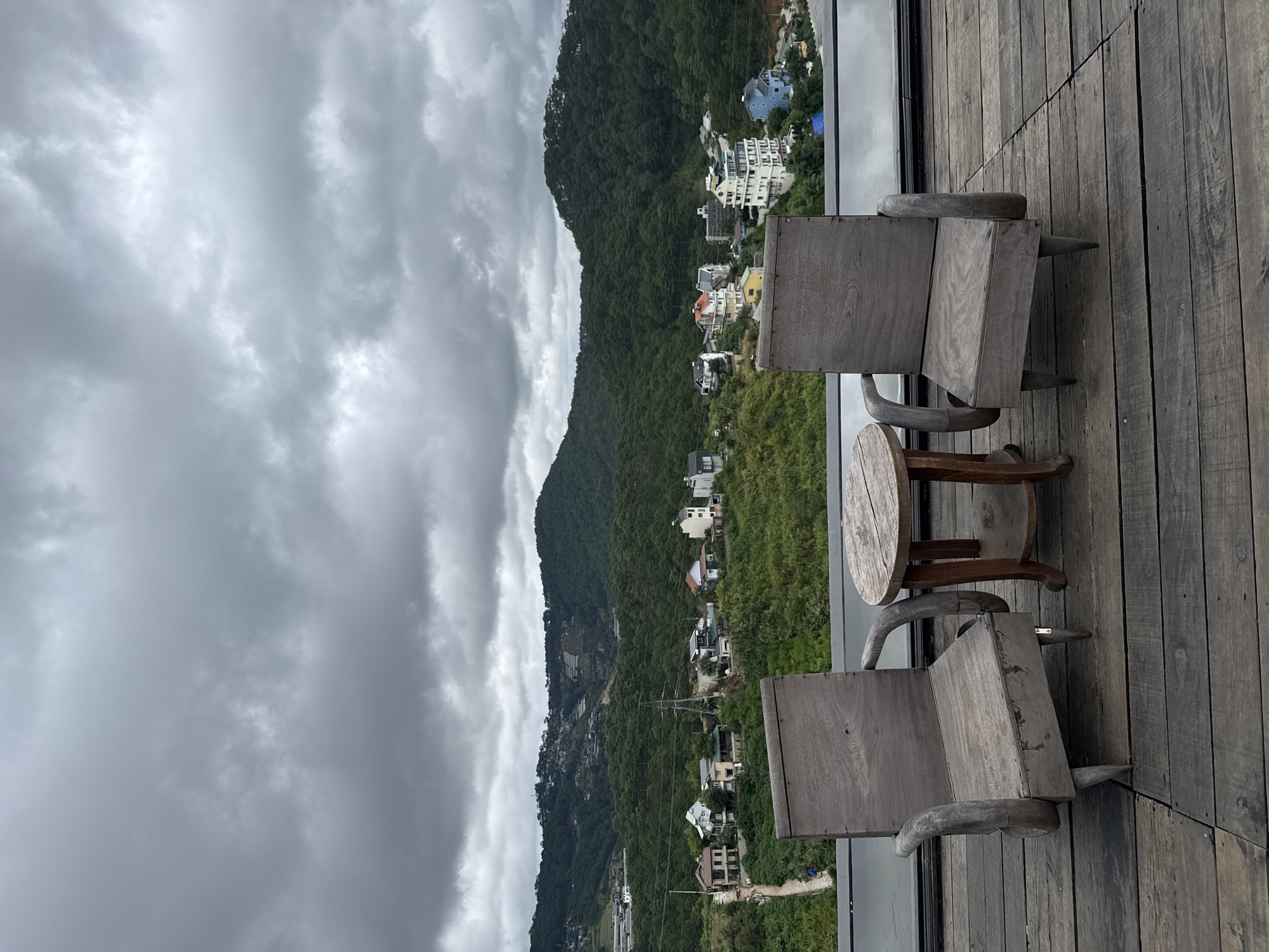 The image captures a scenic view of a mountainous landscape with scattered houses and lush greenery under a cloudy sky. In the foreground, two weathered wooden chairs and a small wooden table are placed on a dark wooden deck. The chairs have a rustic design with curved armrests and square seats. The table is round with a tiered base.
The middle ground features a hillside dotted with various houses, some modern and multi-story, others smaller and more traditional. The houses are surrounded by dense green vegetation, suggesting a natural and somewhat undeveloped area. A winding path is visible leading up to some of the houses.
The background is dominated by rolling hills covered in dark green trees, creating a sense of depth and vastness. The sky is overcast with heavy, grey clouds, casting a diffused light over the entire scene. The overall atmosphere is one of tranquility and natural beauty, with the overcast weather adding a slightly dramatic or moody element. This view is characteristic of the hilly topography and the prevalent residential development found in the outskirts of Da Lat, Vietnam, a city known for its cool climate and picturesque landscapes.