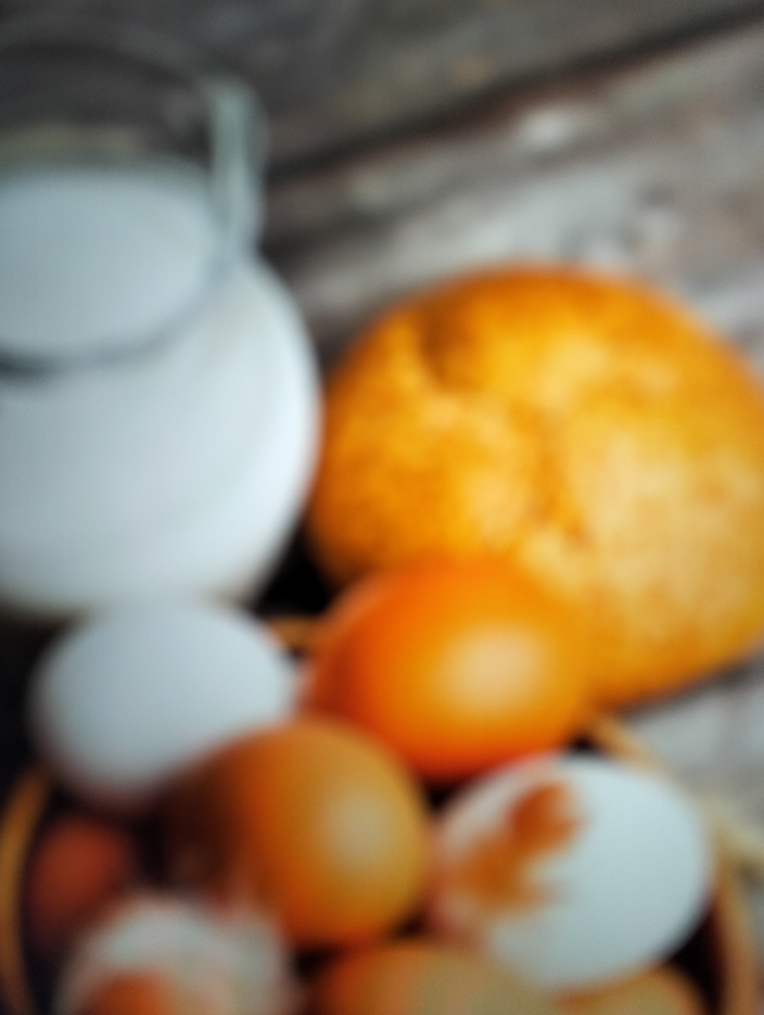 The image shows a collection of food items, including a glass jug filled with a white liquid, likely milk, positioned on the left side. To the right of the jug, a golden-brown loaf of bread is visible, with a rough texture. In the foreground, several eggs are arranged in a basket. Some eggs are white, while others are brown, and one appears to have some speckling. The arrangement suggests a still life of basic food staples. The background is blurred, but appears to be a wooden surface, possibly a table. There are no people or specific location markers in the image that would indicate it is in Payagaboh, Indonesia.