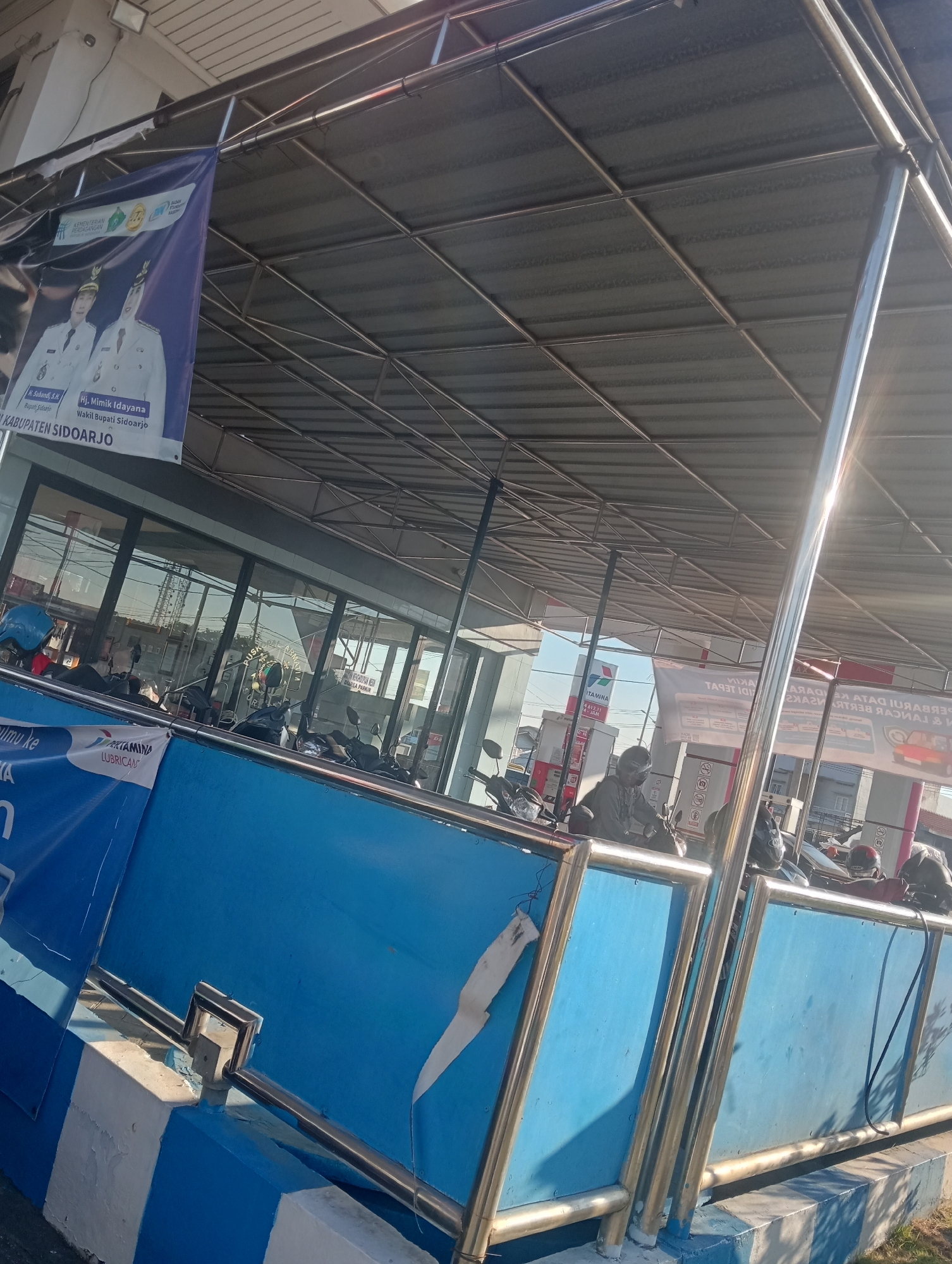 The image captures an outdoor scene at a gas station, likely in Sawotratap, Indonesia. The foreground is dominated by a blue barrier with white stripes on its base, topped with a silver railing. Behind the barrier, several motorcycles are parked under a metal roof supported by columns. Some individuals, wearing helmets, are seated on the motorcycles. The background shows a building with large glass windows, a gas pump, and a banner with images of two uniformed officials and text in Indonesian. Another banner with the Pertamina logo is also visible. The environment appears bright, suggesting it is daytime, with sunlight reflecting off the metal structures.