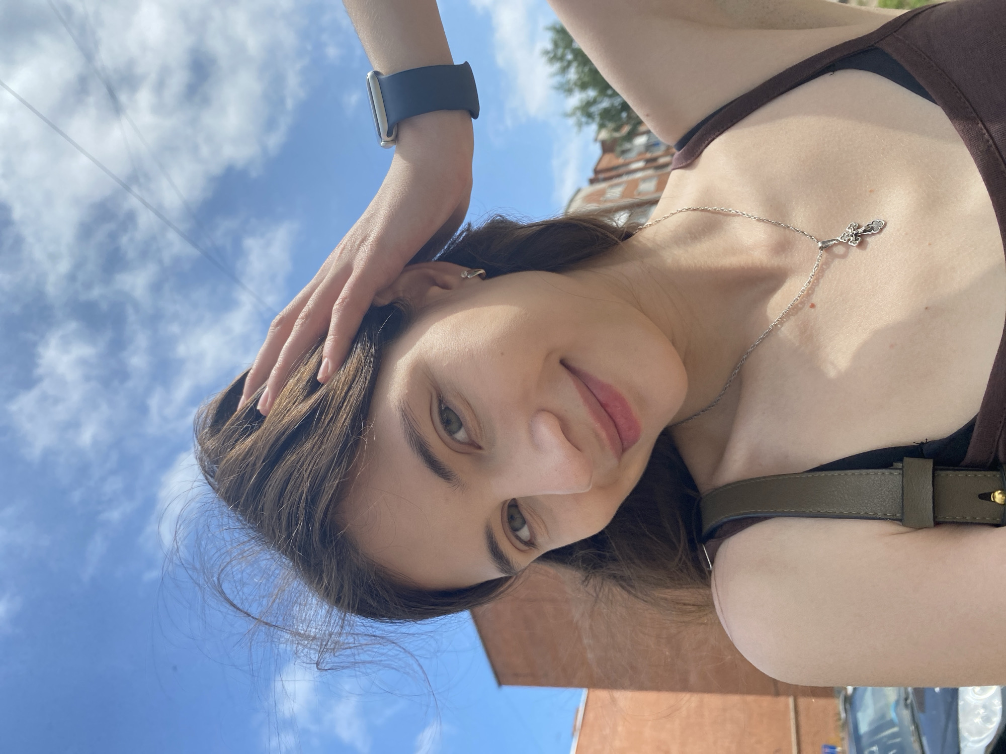 A young woman with long brown hair is taking a selfie outdoors in Votkinsk, Russia, on a sunny day with a blue sky and scattered clouds. She is wearing a brown tank top, a silver necklace with a cross pendant, and a dark smartwatch on her left wrist. Her right hand is in her hair, and her left arm is bent behind her head. In the background, there is a brick building and trees. The photo is taken from the chest up, with the woman's face being the primary focus.