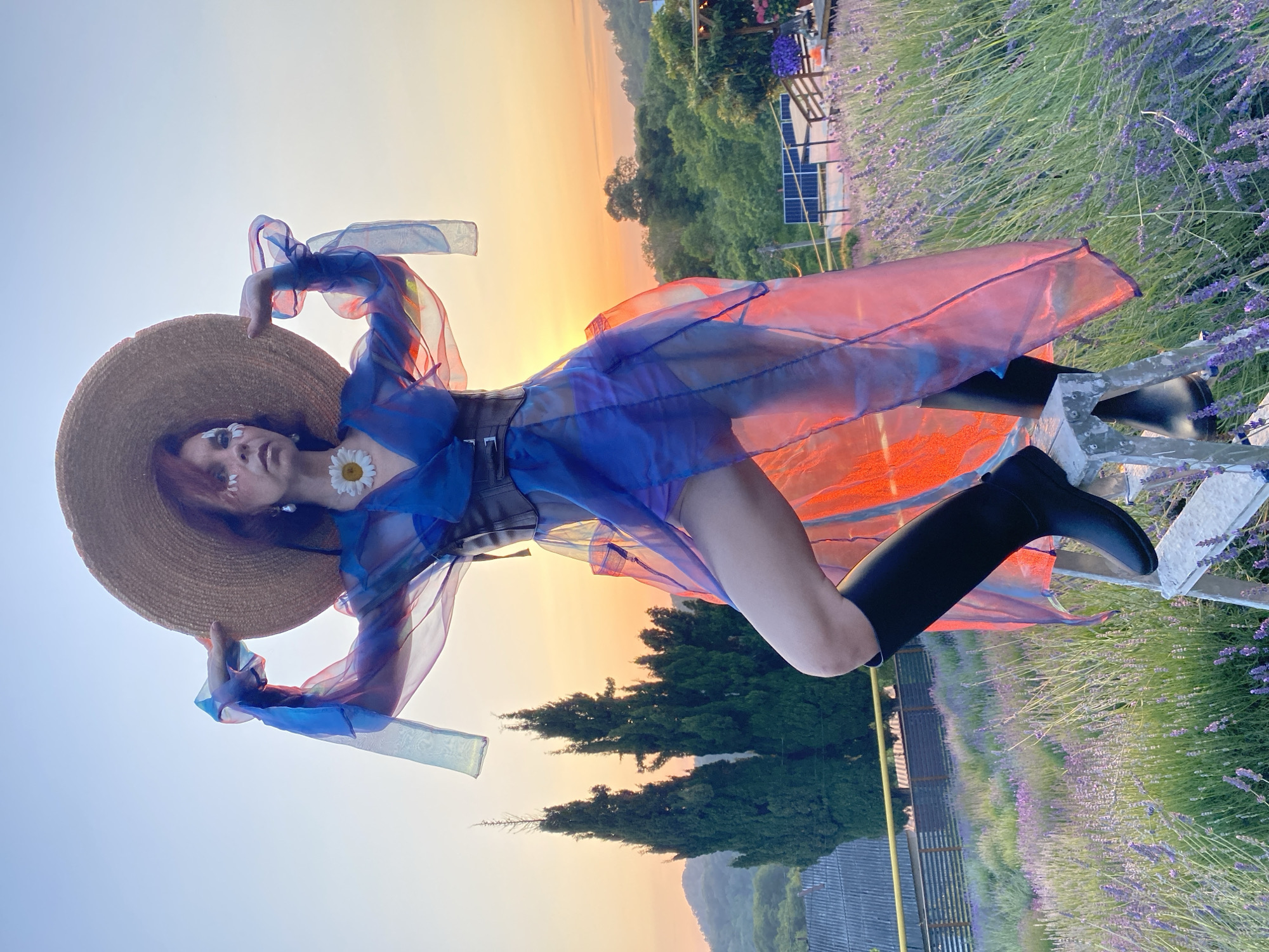 A person stands on a stepladder in a lavender field at sunset. The person is wearing a wide-brimmed straw hat, a blue and pink sheer dress, a brown corset, and black boots. The person's face is painted with white and light purple makeup, and a daisy is attached to their necklace. In the background, there are cypress trees, rolling hills, and a solar panel array. The sky is a gradient of orange, yellow, and blue, indicating the sun is setting. The location is identified as Verkhneye Uchdere, Russia.