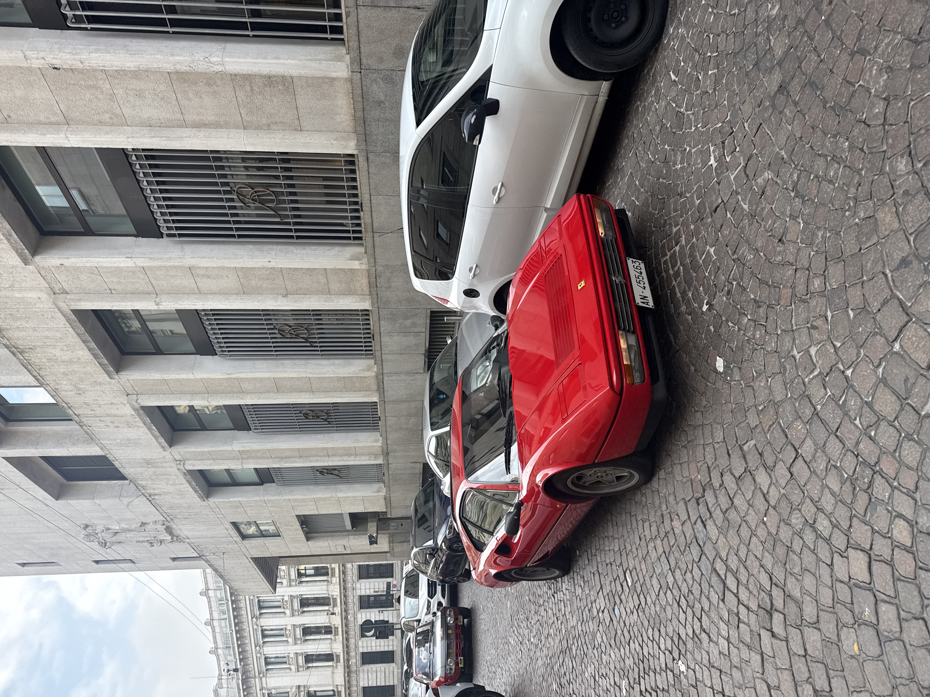 This image depicts an urban street scene in Milan, Italy, specifically in the vicinity of Piazza della Scala. The foreground features an antique cobblestone street.
**Subjects/Objects:**
* **Vehicles:** The prominent subject is a bright red, vintage Ferrari sports car, likely a 308 or 328 model, parked at an angle facing the viewer. It bears an Italian license plate "AN-455463" (Ancona province). To its immediate right, a white compact SUV or hatchback with black wheels is parked parallel to the curb. Further back, several other vehicles are visible, including a dark blue SUV, a red and black vintage Lancia Fulvia Coupé on the far left, and a white Ford Kuga SUV. The street appears congested with parked or stationary vehicles.
* **Buildings:**
* **Right:** A multi-story, modern building with a grey, textured stone facade dominates the right side. This building, identified as the former Banca Commerciale Italiana headquarters, features recessed rectangular windows, some protected by vertical metal grilles. Two prominent relief sculptures depicting human figures (possibly classical or allegorical) are visible on the upper levels. Below these, stylized 'B' monograms are integrated into the window grilles.
* **Left Background:** A grand, ornate, classical-style building with multiple stories, light-colored stone, numerous large windows, pilasters, and decorative elements is partially visible behind other vehicles. A partial "UNI" sign is discernible on this structure.
* **Street Furniture:** A black circular sign with a white crescent moon shape is visible on the left side of the street, and a streetlight pole is present.
* **People:** One individual is partially visible in the mid-background behind the white Ford Kuga, obscured by the vehicle.
**Environment:**
The sky is overcast and uniformly grey, indicating diffused daylight. The street is paved with dark grey and brown cobblestones, showing minor wear and some debris. A narrow strip of smoother pavement or asphalt runs alongside the building on the right.
**Actions/Interactions:**
The scene is static, primarily showing parked or stationary vehicles. No overt actions or interactions between individuals are discernible.