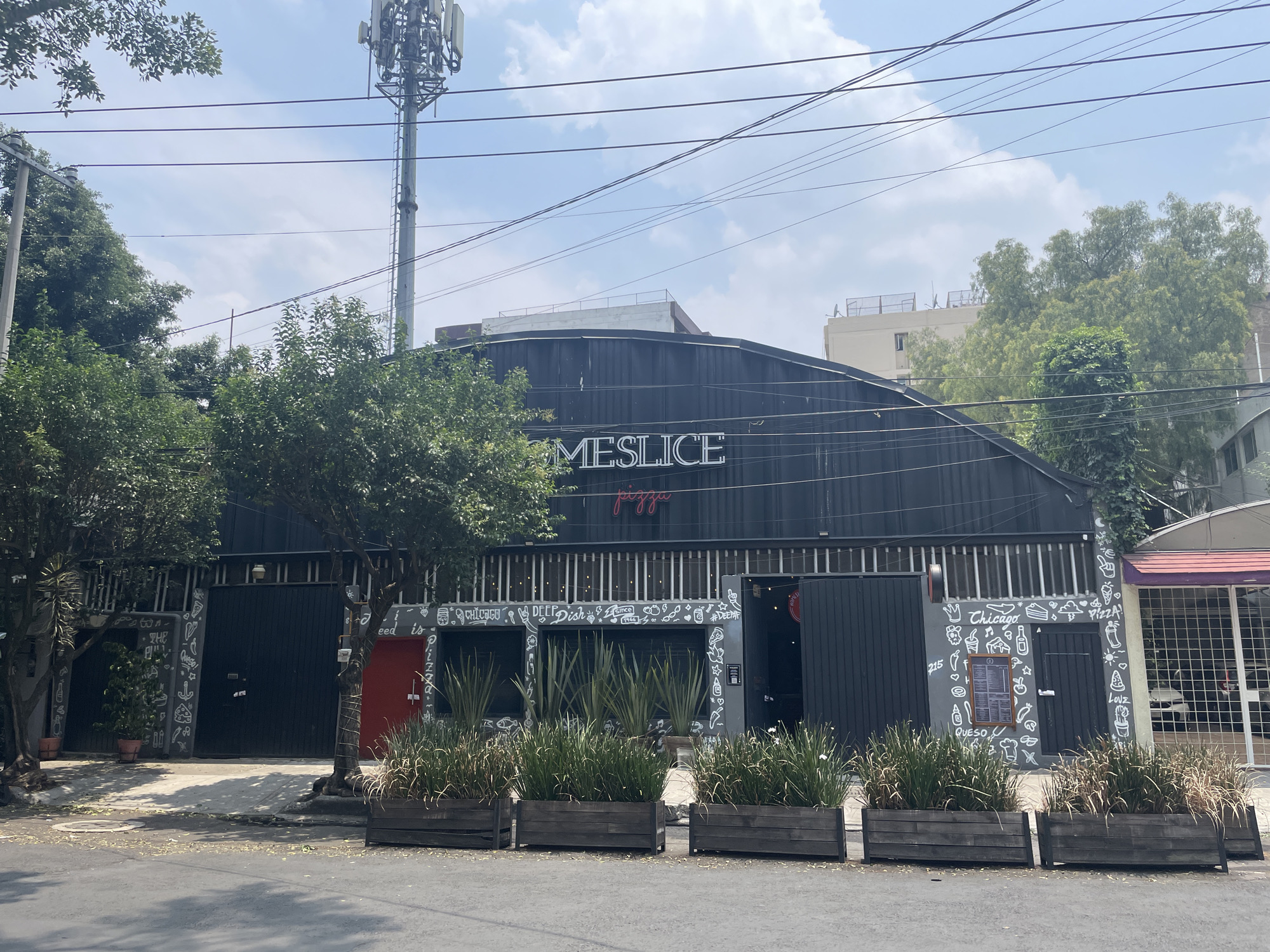 The image depicts the exterior of "Meslice Pizza", located in Mexico City, Mexico. The restaurant has a dark gray facade with a curved roof. A sign reading "MESLICE pizza" is prominently displayed above the entrance. The facade is decorated with white drawings. Planter boxes containing plants line the front of the restaurant. A cell tower is visible behind the building. Overhead power lines stretch across the sky. A street runs in front of the restaurant, and trees are visible on either side of the building.