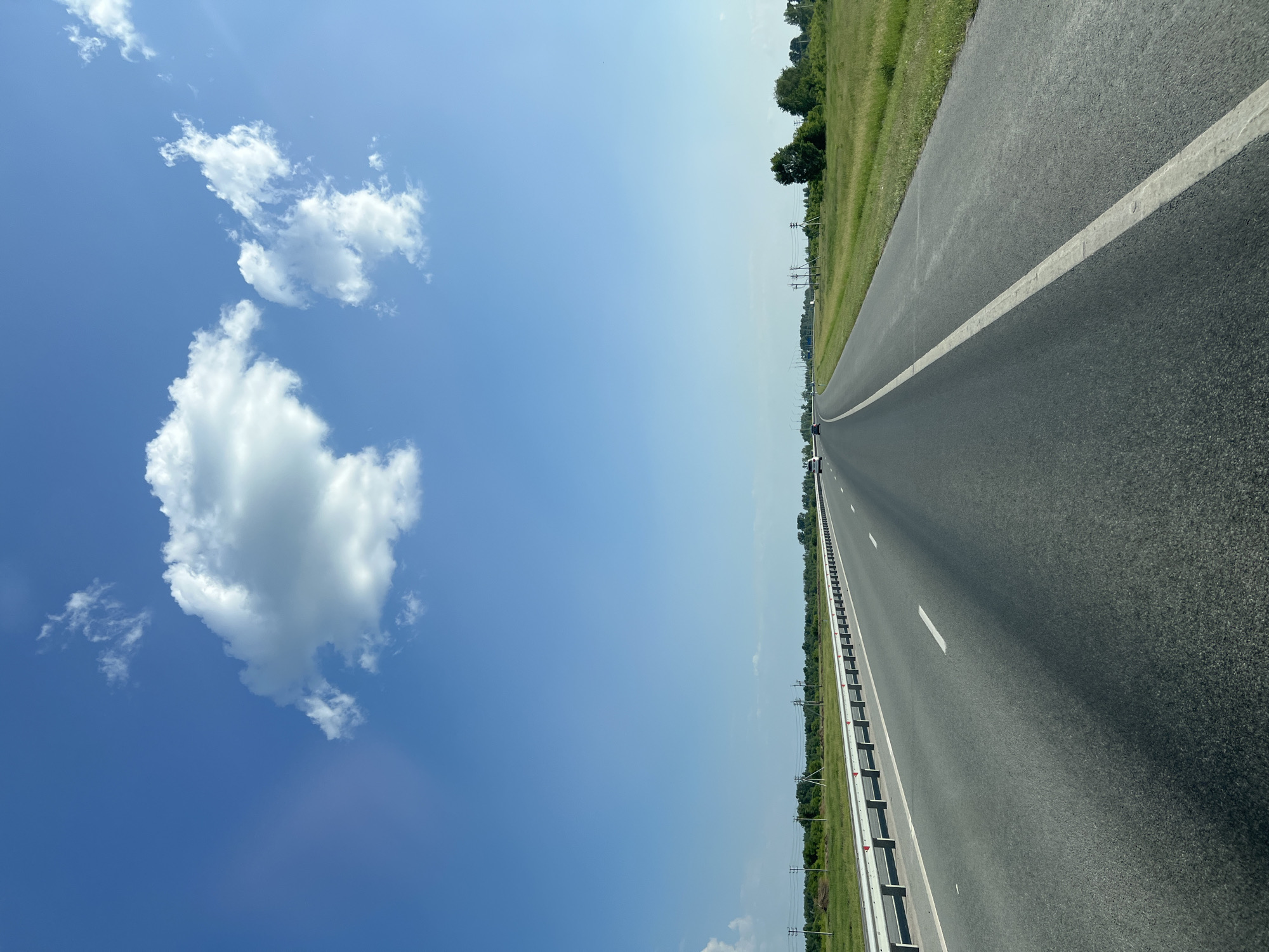 The image depicts a view from a vehicle traveling on a multi-lane highway in Verkh-Tula, Russia. The road is asphalt, with clear white lane markings. A metal guardrail runs along the left side of the highway, separating it from a grassy verge. Beyond the verge, there are fields, trees, and utility poles. The sky is a clear blue with scattered white clouds. In the distance, a few vehicles can be seen on the highway. The overall impression is of a bright, sunny day on a rural highway.