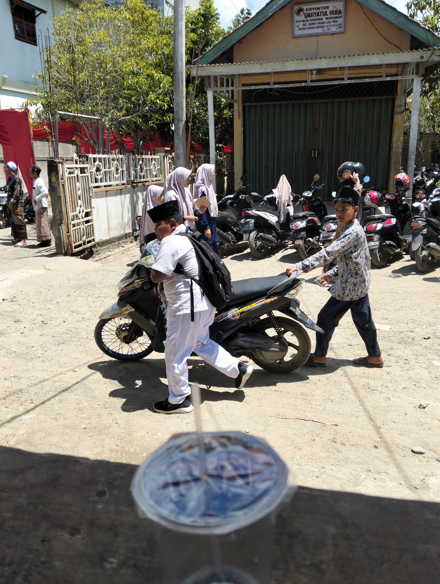 In Teungoh, Indonesia, a sunny outdoor scene unfolds in front of a building with a sign identifying it as "KOPONTREN QARYATUL HUDA". The building has a light brown facade and a green roll-up door. Several motorcycles are parked in front of the building. In the foreground, a boy wearing a patterned shirt and dark pants walks away from the viewer, holding a drink with a straw in his right hand. To the left of him, another person in white attire, possibly a student, walks towards a motorcycle, carrying a backpack. Several other individuals are visible in the background, including some wearing headscarves, and a red fabric is draped on a fence to the left of the building. The ground is dirt and gravel.
