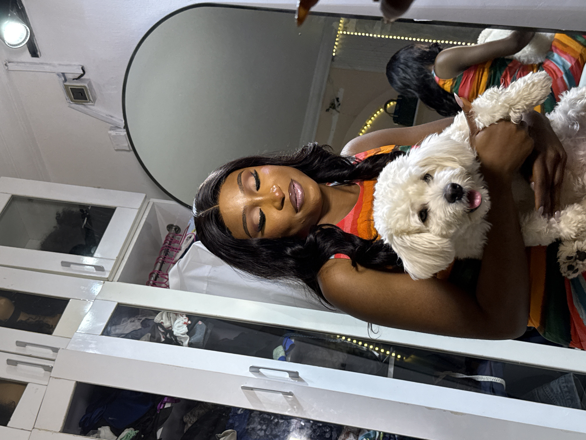 The image depicts a woman holding a white poodle-mix dog in what appears to be a room in Abuja, Nigeria. The woman is positioned centrally, with her eyes closed and a gentle smile, and is dressed in a colorful, striped dress. The dog, held securely in her arms, is looking towards the camera with its tongue slightly out. Behind the woman, a large, arched mirror reflects part of the room, showing another woman in a similar dress and the dog. The environment includes a white wardrobe with glass doors revealing clothing and personal items, a wall with a light fixture and an electrical socket, and a string of decorative lights. The overall lighting suggests an indoor setting, possibly a bedroom or dressing area.