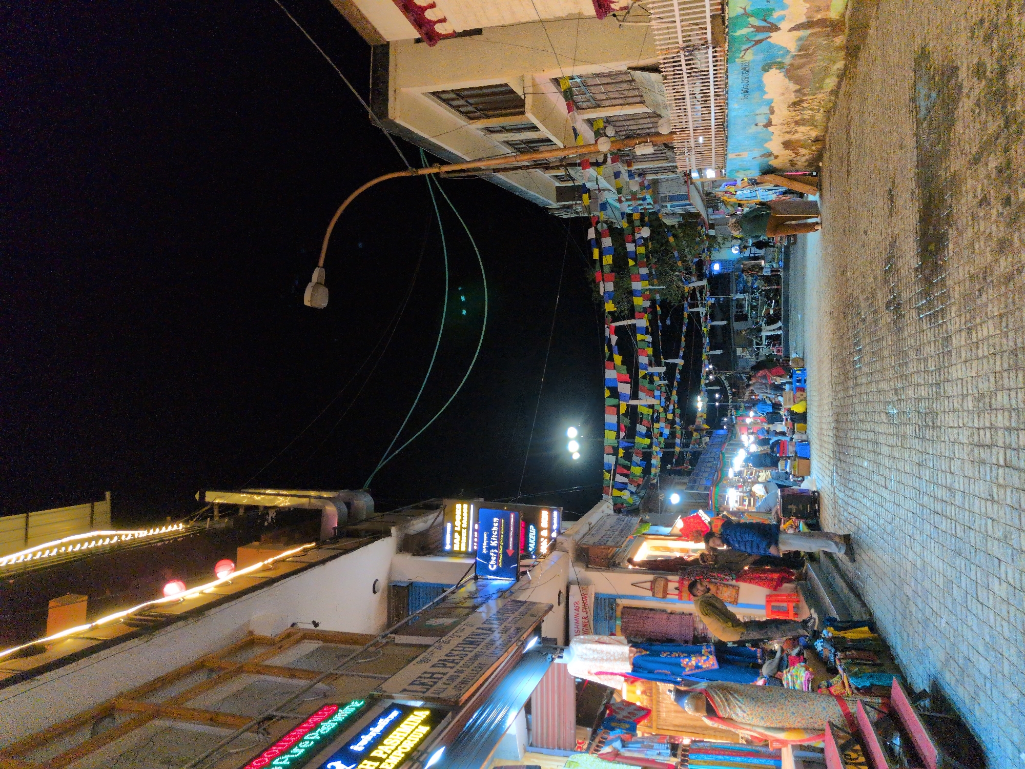 This is a nighttime outdoor shot of a street market in Leh, India. The scene is bustling with activity, illuminated by streetlights, shop signs, and strings of decorative lights. Several people are present, browsing goods and walking along the paved walkway.
Shops line the street, displaying a variety of items, including textiles, clothing, and possibly souvenirs. Signs for "Chef's Kitchen" and "Leh Pashmina Emporium" are clearly visible, indicating food and local crafts are available. Prayer flags, commonly seen in Buddhist regions, are strung across the street, adding a touch of cultural detail. The overall atmosphere appears lively and vibrant, typical of a market in the evening. The ground appears to be wet, suggesting recent rain or damp conditions. The sky is dark, confirming it is nighttime.