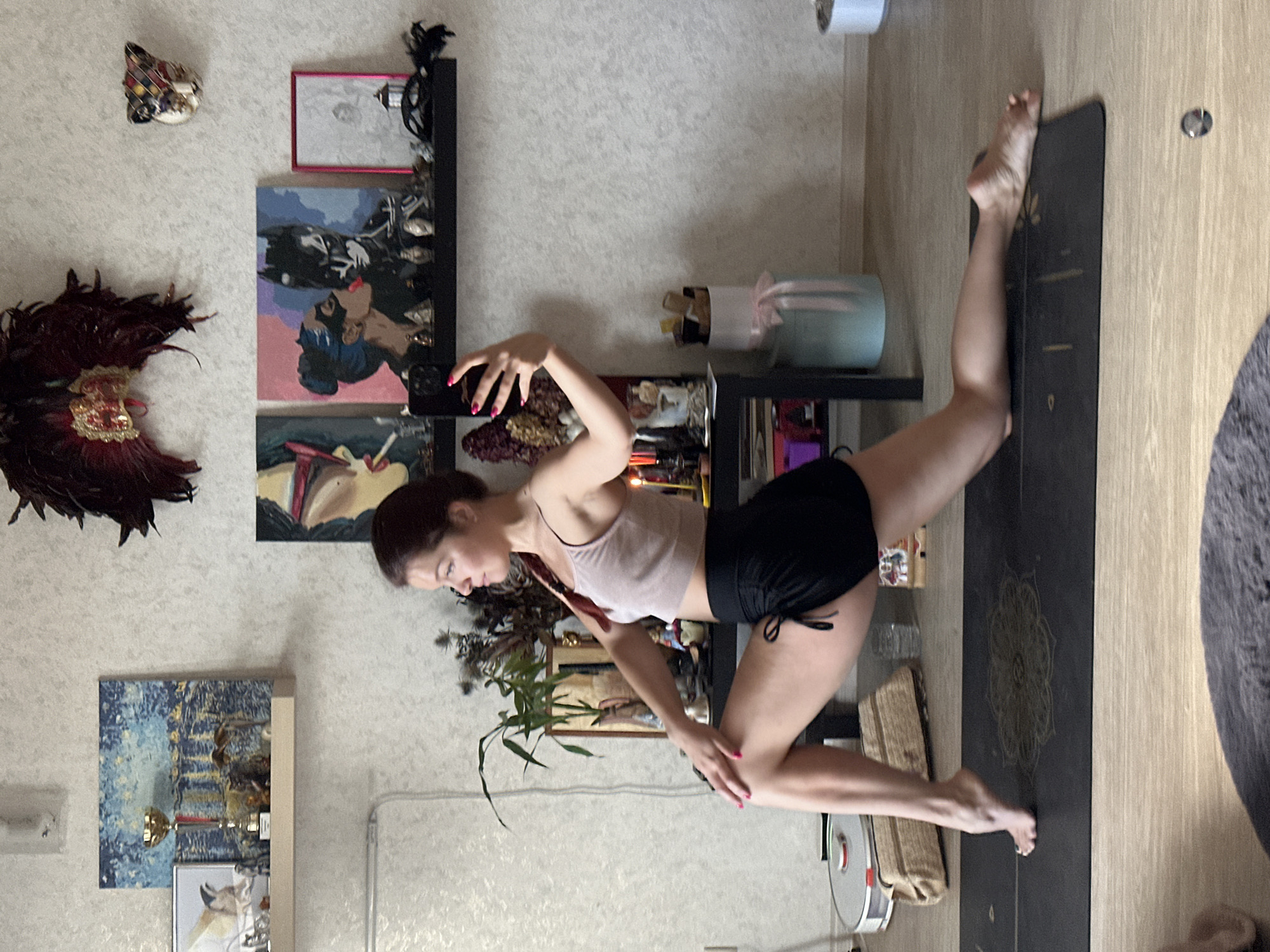 A young woman with dark hair is performing a yoga pose on a mat in a living room. She is wearing a light-colored crop top and black shorts. The room is decorated with several pieces of art on the wall and shelves, including paintings and a mask. There are also plants, decorative items, and what appears to be a robot vacuum cleaner in the background. The floor is wooden. The specific location in Vladimir, Russia, is not discernible from the image.