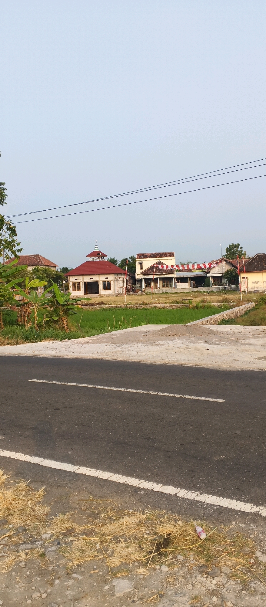 In Japanan, Indonesia, a village scene unfolds under a pale sky. A paved road with a white dashed line runs across the foreground. The roadside is bordered by dry, yellowish grass and scattered small rocks. Beyond the road, a patch of green rice paddy fields stretches towards a cluster of buildings.
In the middle ground, several structures are visible. The most prominent is a building with a red roof and white walls, possibly a mosque or community hall, featuring arched windows and a decorative dome. To its right, a two-story building with a white facade and a tiled roof stands, adorned with red and white striped banners. Further to the right, another building with a dark tiled roof is partially visible. These buildings appear to be in various states of construction or maintenance.
In the background, more buildings and trees can be seen, suggesting a residential or developed area within the village. Power lines stretch across the upper portion of the image. The overall impression is a quiet, rural setting in a developing Indonesian village. No people are visible in the scene.