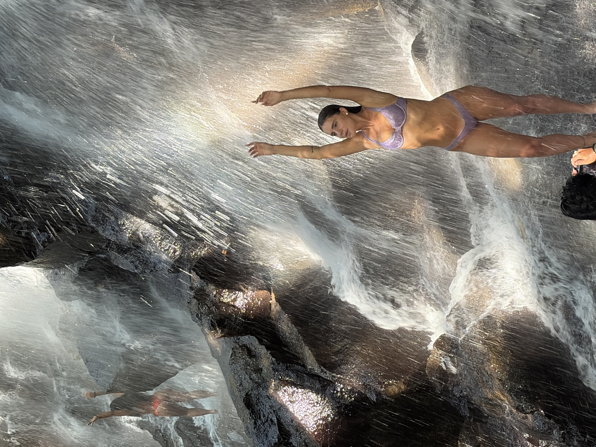 The image depicts a natural scene with a waterfall cascading over rocks. In the foreground, a woman in a light purple bikini stands with her arms raised, facing the waterfall. To the left and further back, another person in a red bikini stands with their arms extended. The environment is characterized by flowing water, wet rocks, and dappled sunlight creating a shimmering effect. The presence of a waterfall suggests an outdoor, natural setting, possibly a tourist attraction or a secluded spot for recreation. The location is identified as being in the city of Gianyar, Indonesia, known for its beautiful waterfalls and lush landscapes.
