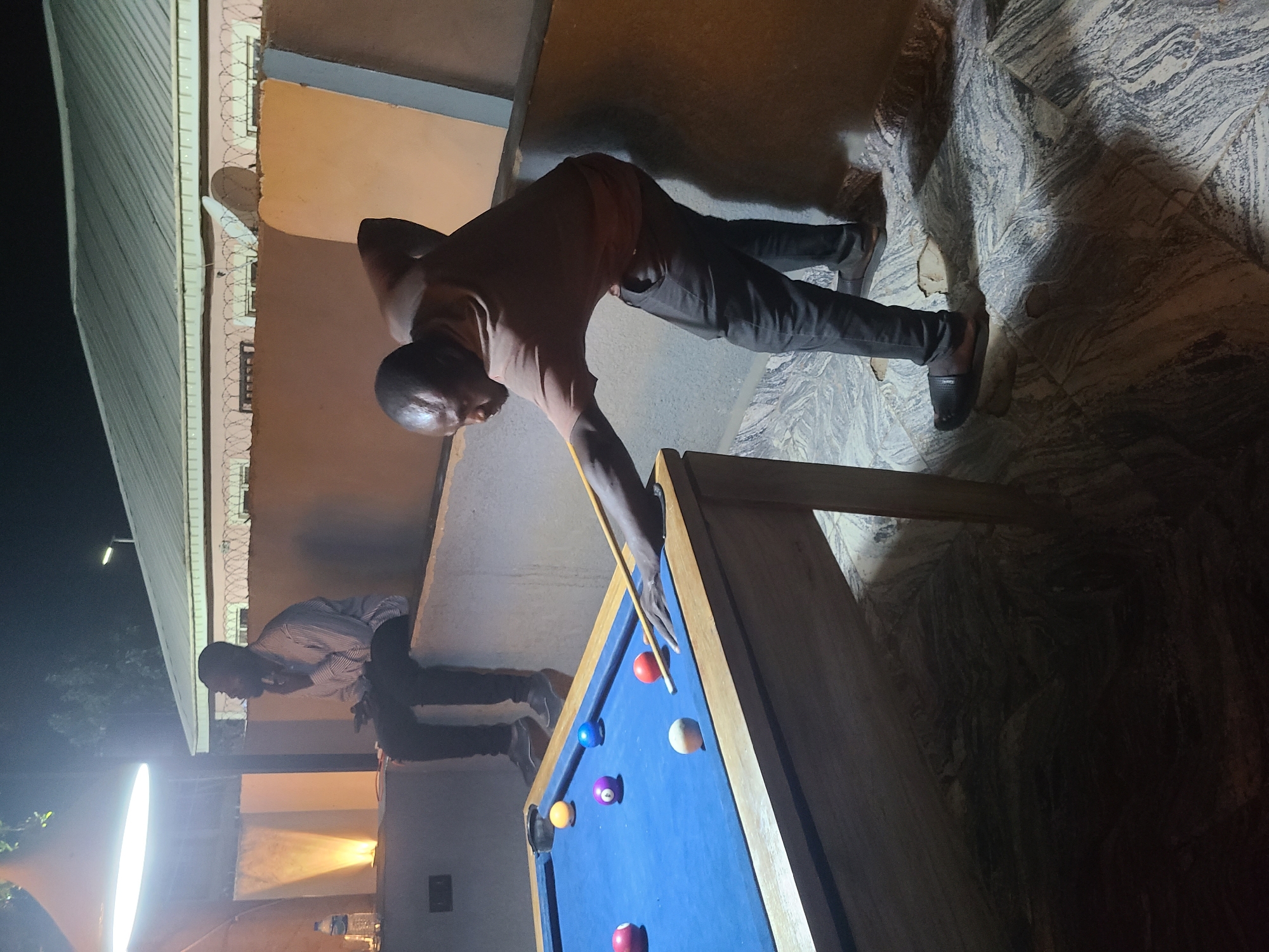 The image depicts a nighttime scene in Gura Top, Nigeria, focusing on two men engaged in a game of pool. The central figure is a man, dressed in a brown shirt and dark trousers, leaning over a blue-felt pool table, meticulously aiming a shot with a cue stick. Several colored pool balls, including red, blue, yellow, and purple, along with a white cue ball, are positioned on the table.
In the background, another man in a striped shirt and dark trousers is visible, appearing to observe the game. The setting is an outdoor or semi-outdoor area, illuminated by artificial light sources; a bright, elongated light source is prominent on the left, while softer lighting glows from structures in the distance.
Architectural elements include a building with a corrugated roof, and a wall or barrier topped with barbed wire, suggesting a secure or enclosed space. A small satellite dish is mounted on a structure in the background. The ground beneath the players features a distinctive, patterned tile or stone surface. A water bottle can be seen on a surface in the background. The overall impression is of a casual, evening leisure activity. No discernible text is visible in the media file.