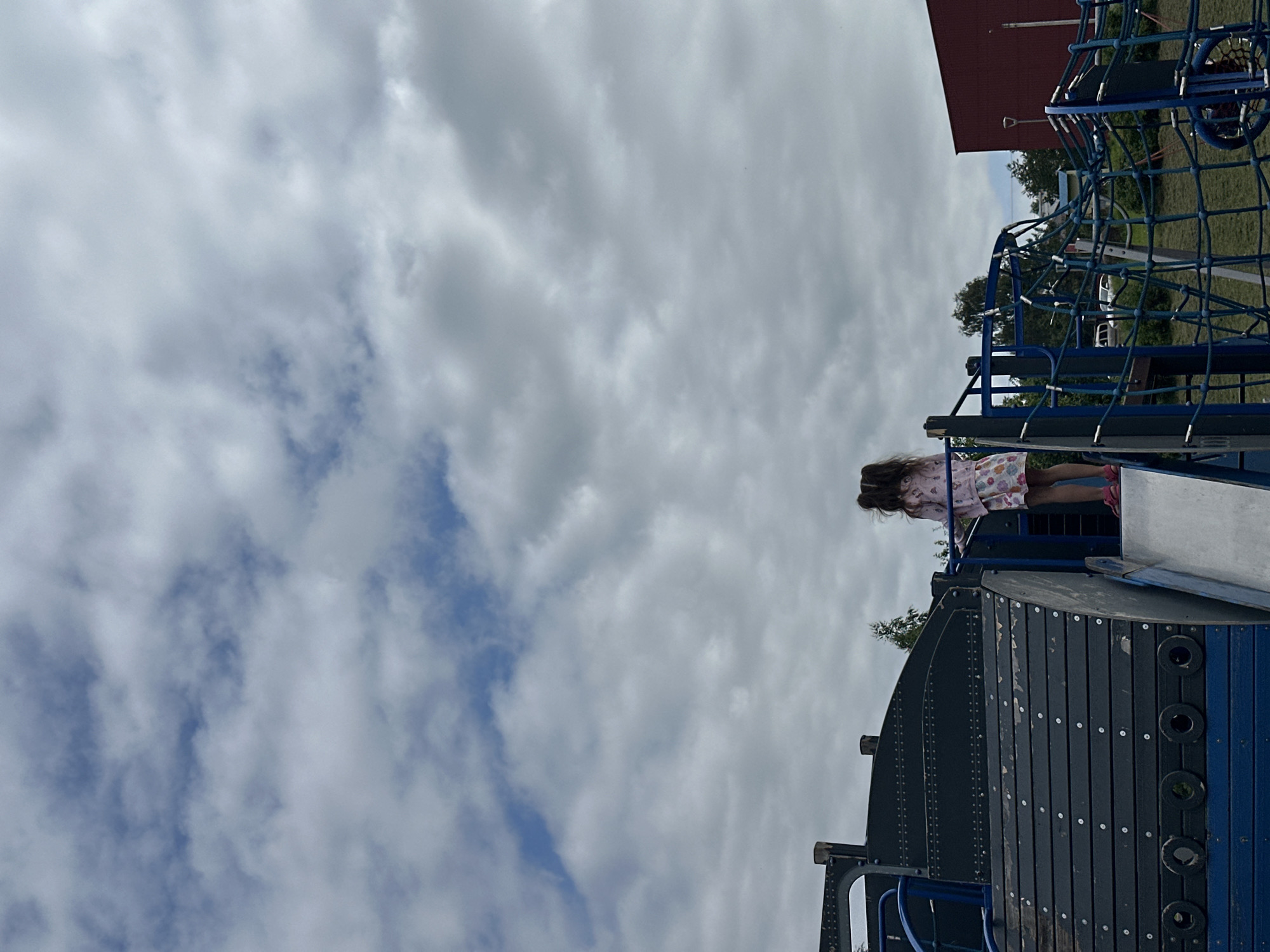 The image shows a young girl standing on a playground structure in Slantsy, Russia. The playground appears to be made of wood and metal, with a blue slide and a rope climbing net. The girl is wearing a pink dress and pink sandals. She is looking away from the camera, towards the sky. The sky is overcast with white clouds. In the background, there is a red brick building and some trees. The overall impression is one of a cloudy day in a playground.